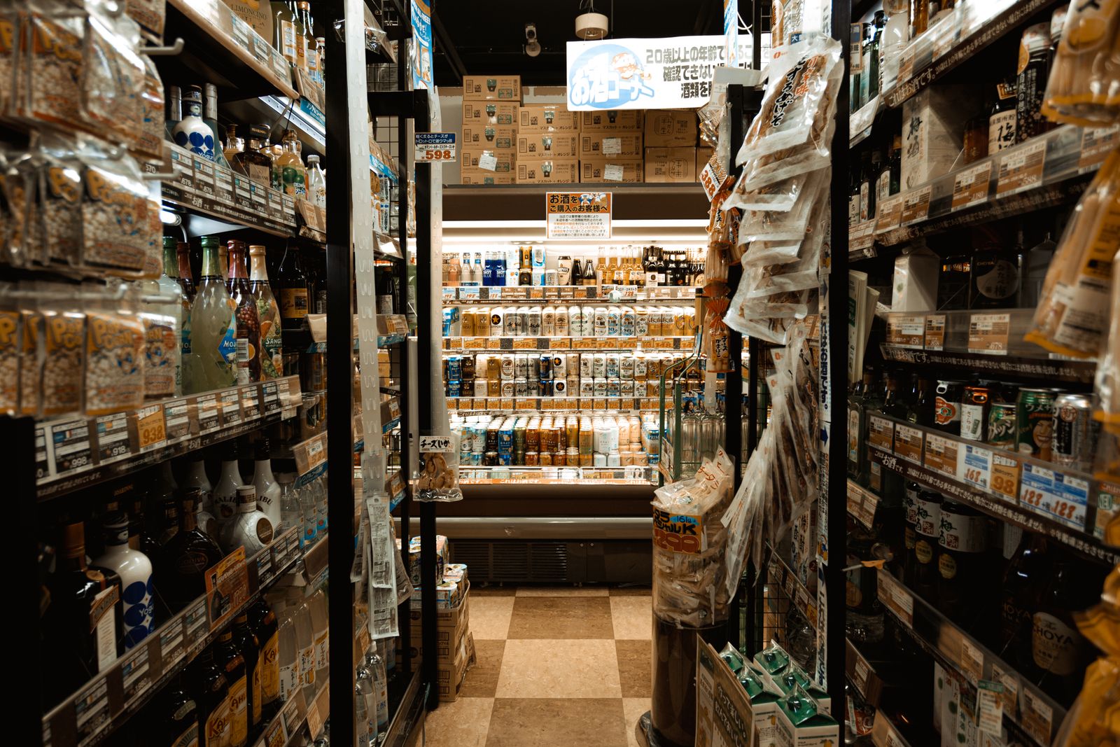 A narrow Japanese drinks aisle with an age-verification notice hanging overhead and shelf tags in Japanese.