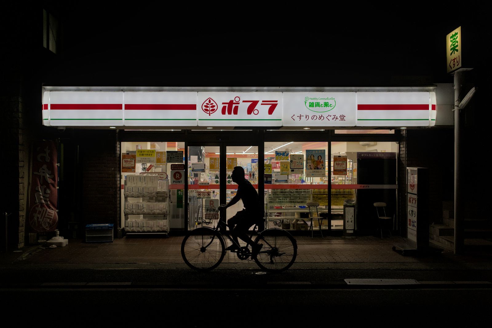 A cyclist in silhouette passing a Poplar convenience store at night in Kyoto, a pharmacy sign glowing above it.