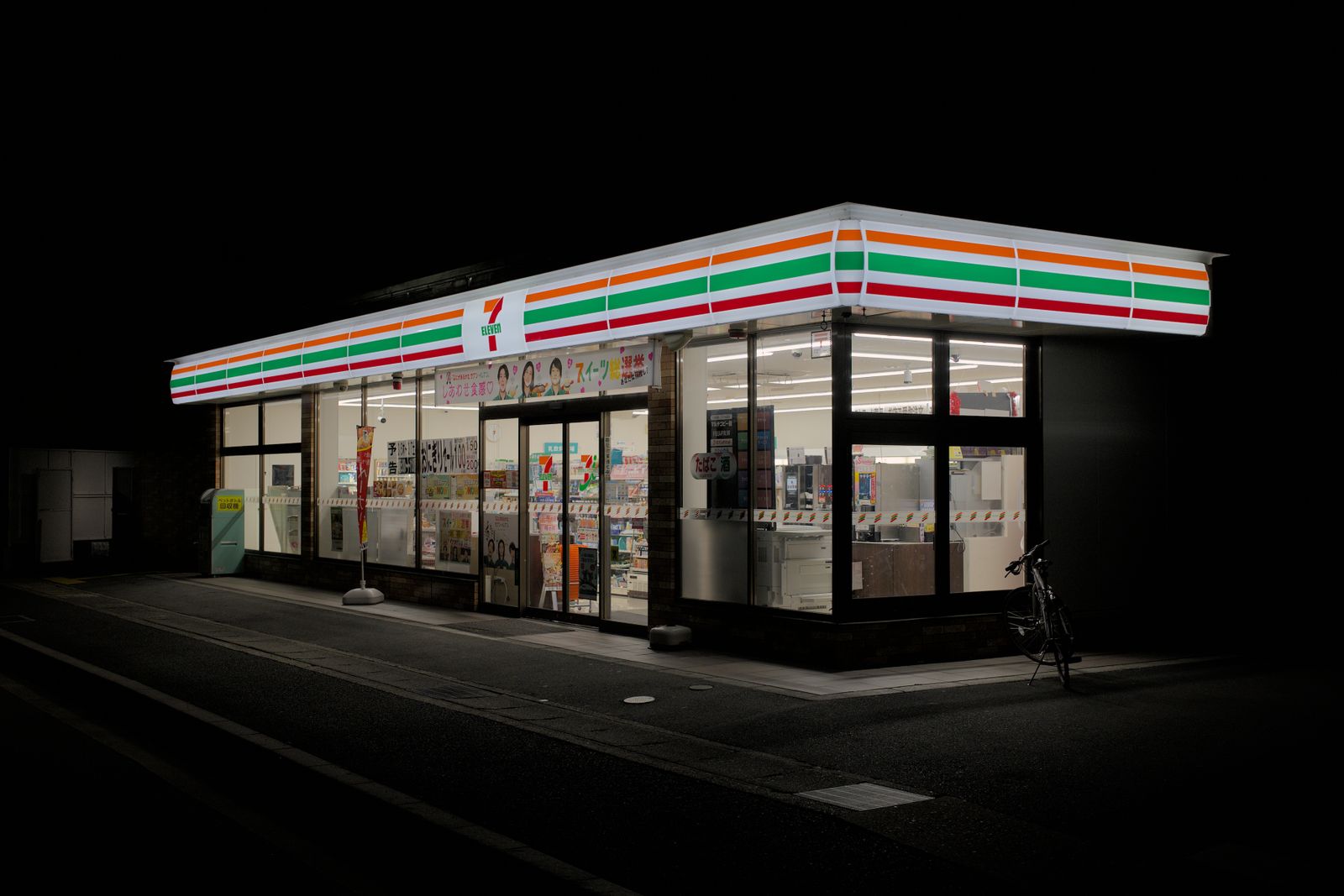 A whole convenience store lit up on an unlit road at night, a photocopier visible through the right-hand window.