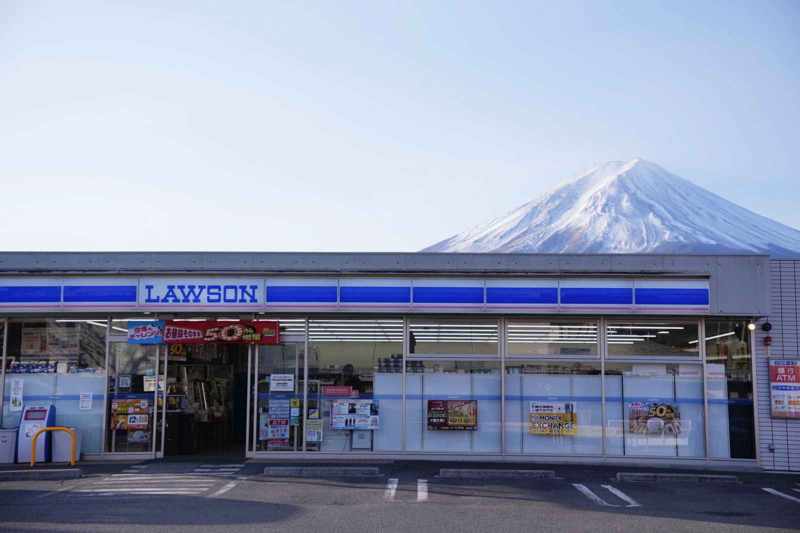 A Lawson storefront in Yamanashi in clear daylight with Mount Fuji rising behind it, an ATM notice and a money-exchange banner on the glass.
