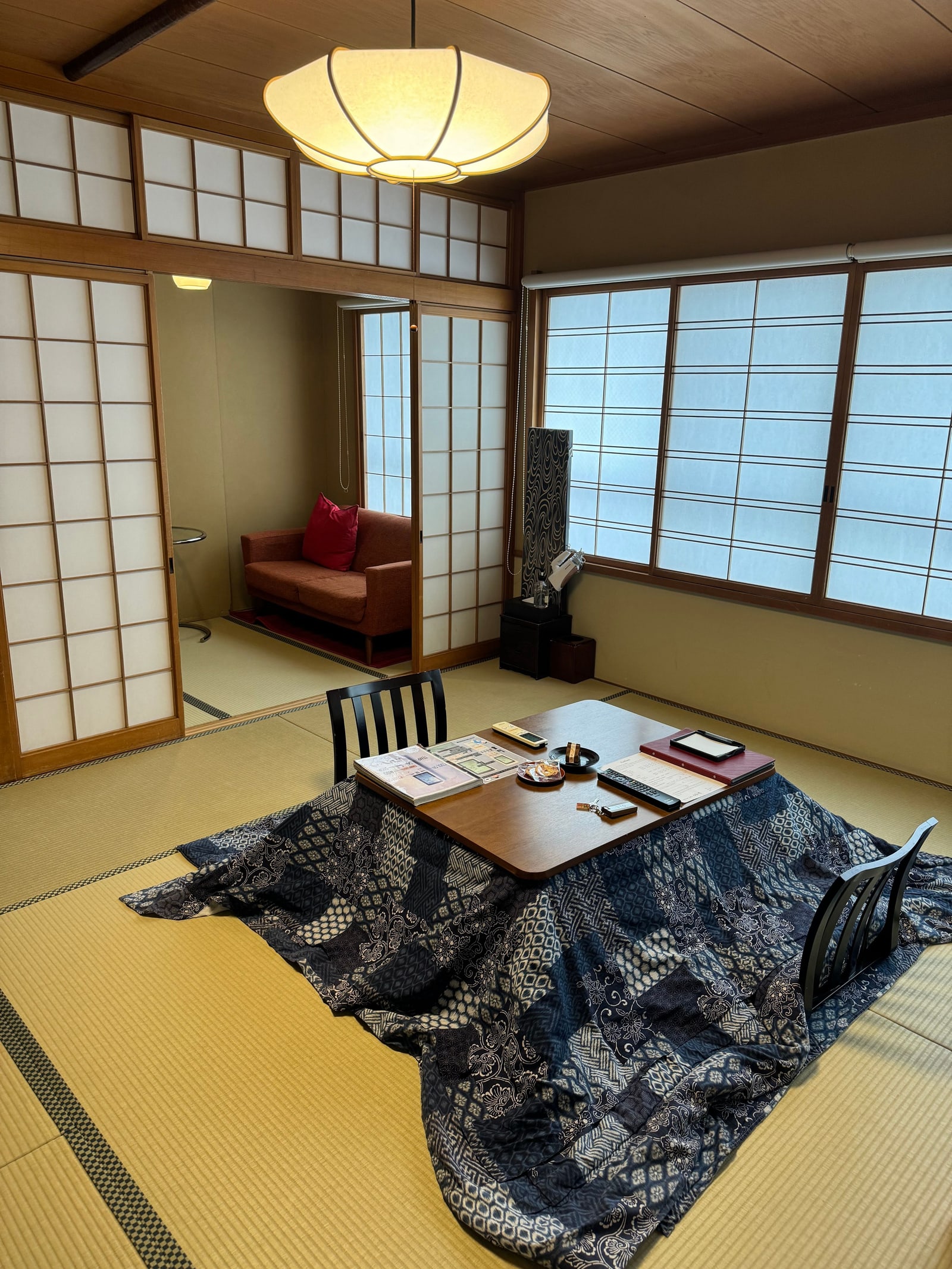 A kotatsu — a low wooden table top laid over a navy patterned quilt — on tatami in a Japanese room with shoji screens, a paper lantern and two low chairs.