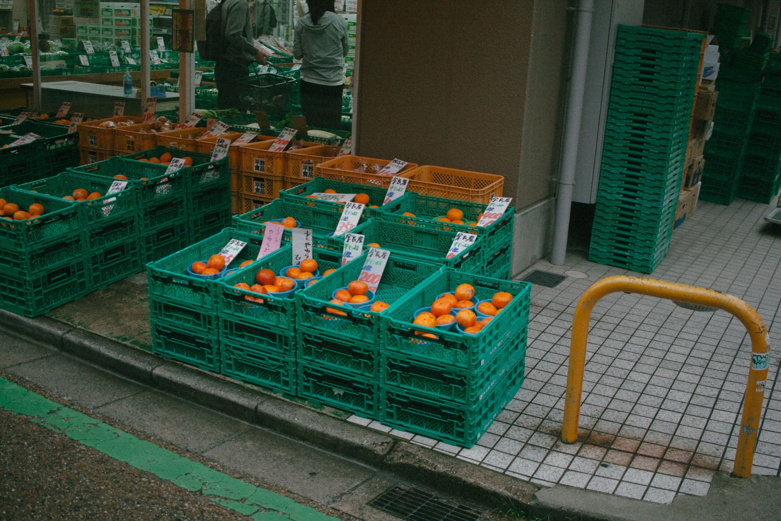 Green and orange plastic crates of mikan with handwritten price tags stacked outside a Japanese greengrocer, two shoppers inside.