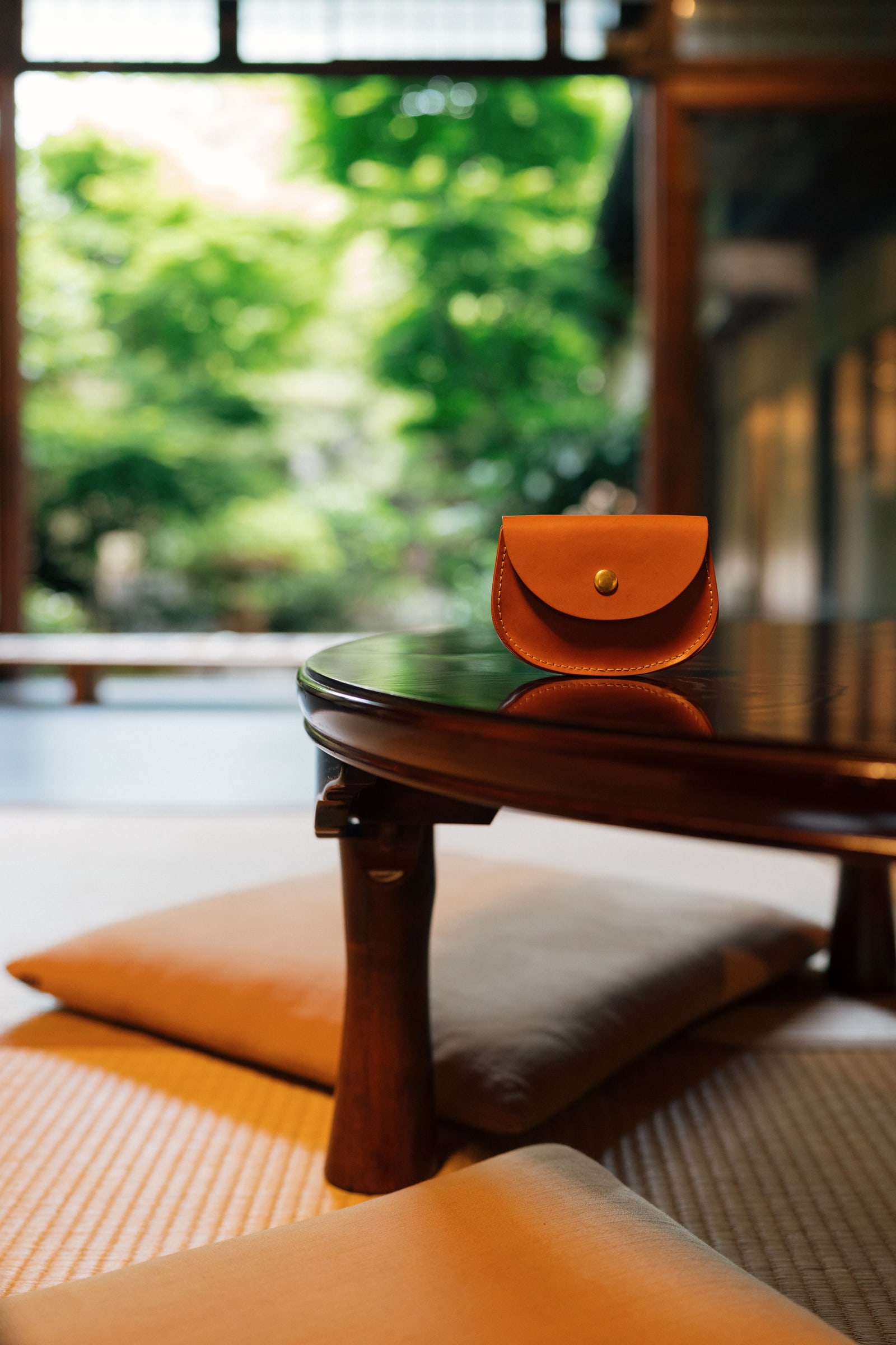 A small round lacquered low table on tatami with floor cushions and a green garden beyond, an orange leather coin purse resting on the table top.