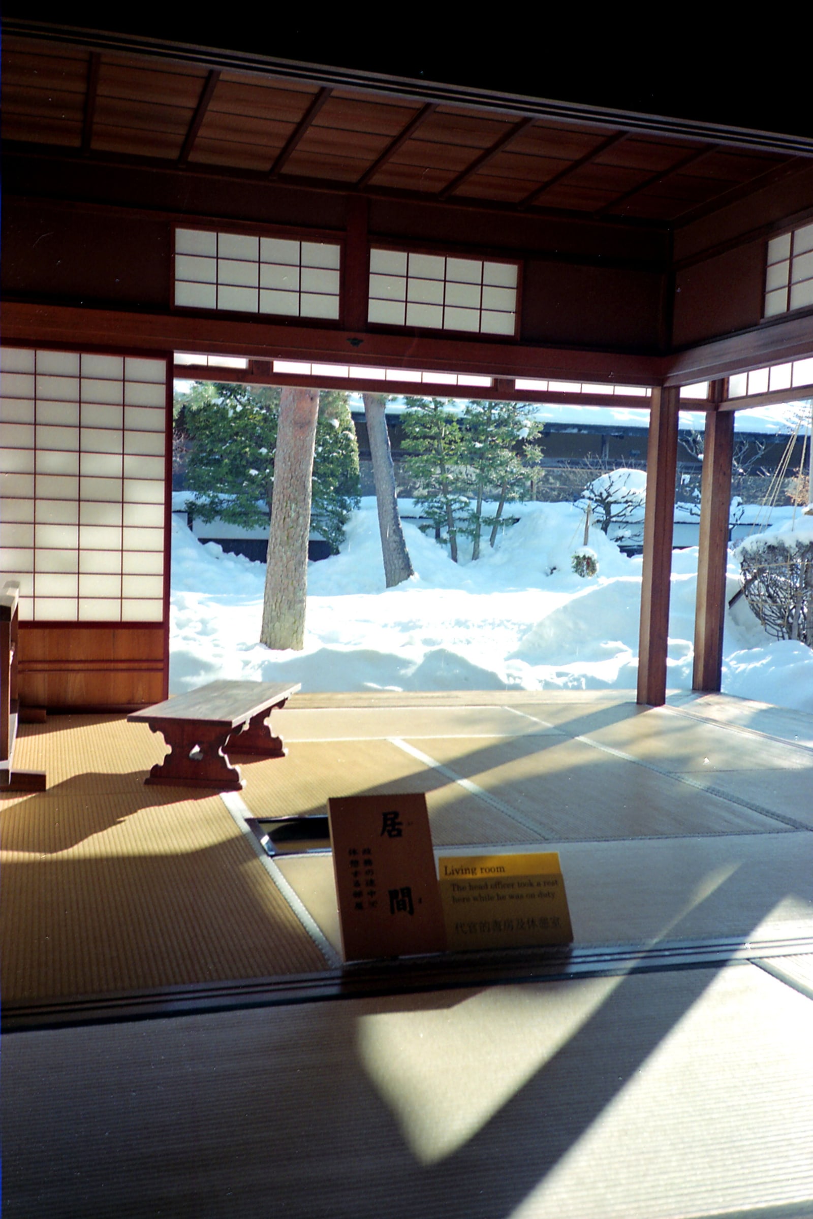 A sunlit tatami room in a historic building in Takayama, open to a snow-covered garden with pines; a low wooden bench-table stands on the tatami and a museum label marks the room as a Living room.