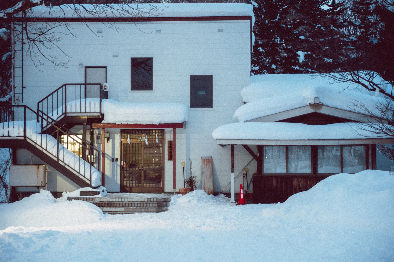 A white two-storey house in Hokkaido at dusk, deep snow on the ground and porch roof, an exterior staircase and a lattice entrance door with a New Year rope decoration.