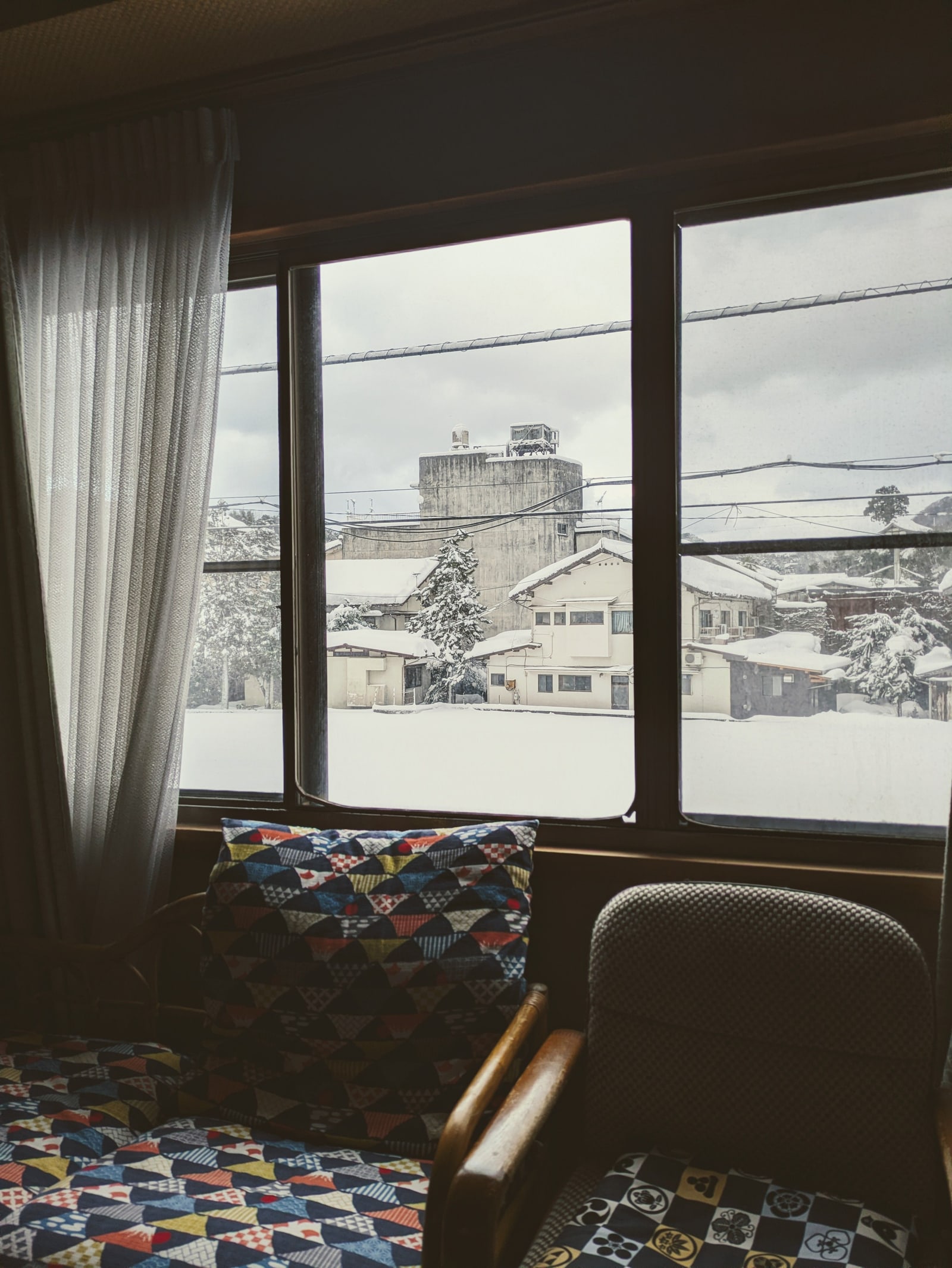 Snow-covered houses and a bare tree seen through a window, with a sheer curtain and two armchairs with patterned cushions in the foreground.
