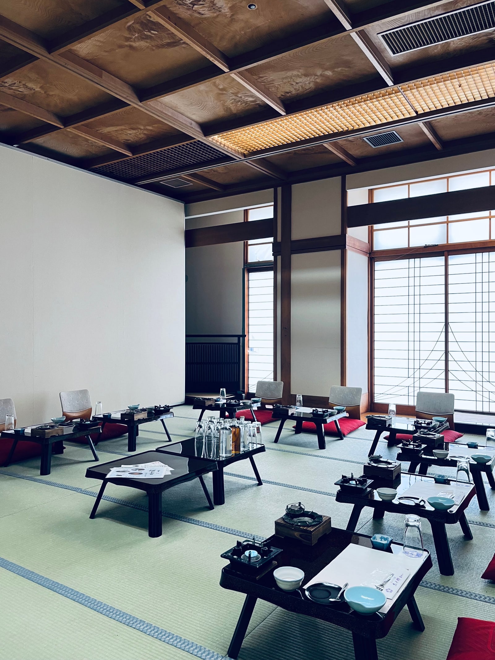 A large tatami banquet room with low lacquered tables, tabletop cookers, and legless floor chairs with red cushions under a coffered wooden ceiling.