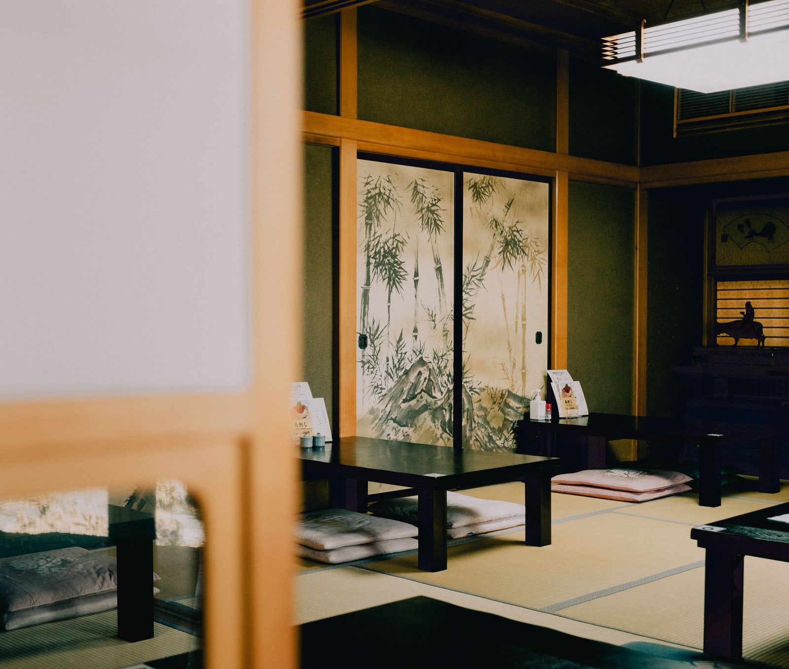 Dark low wooden tables and floor cushions on tatami, seen past a shoji edge, with sliding doors painted with bamboo.