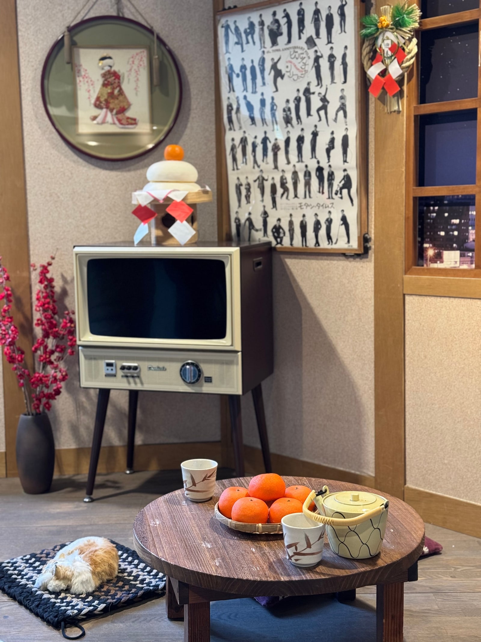 A round low wooden table holding a basket of mikan, a teapot and two cups, in a Japanese-style room with a vintage television, New Year decorations and a plush cat on a mat.