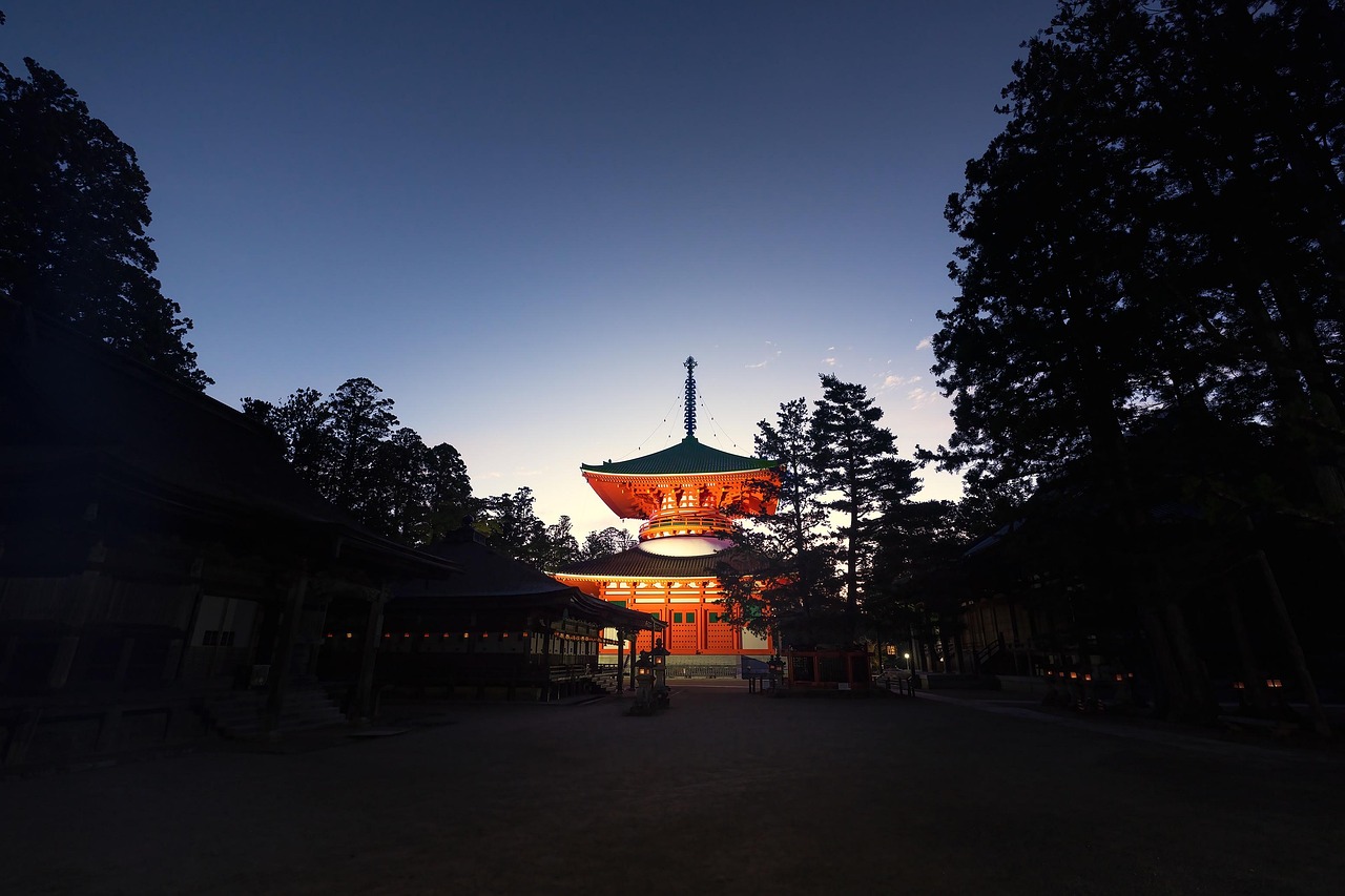 A lit vermilion two-story pagoda at the far end of a dark open precinct framed by cedars, under a deep blue dusk sky.