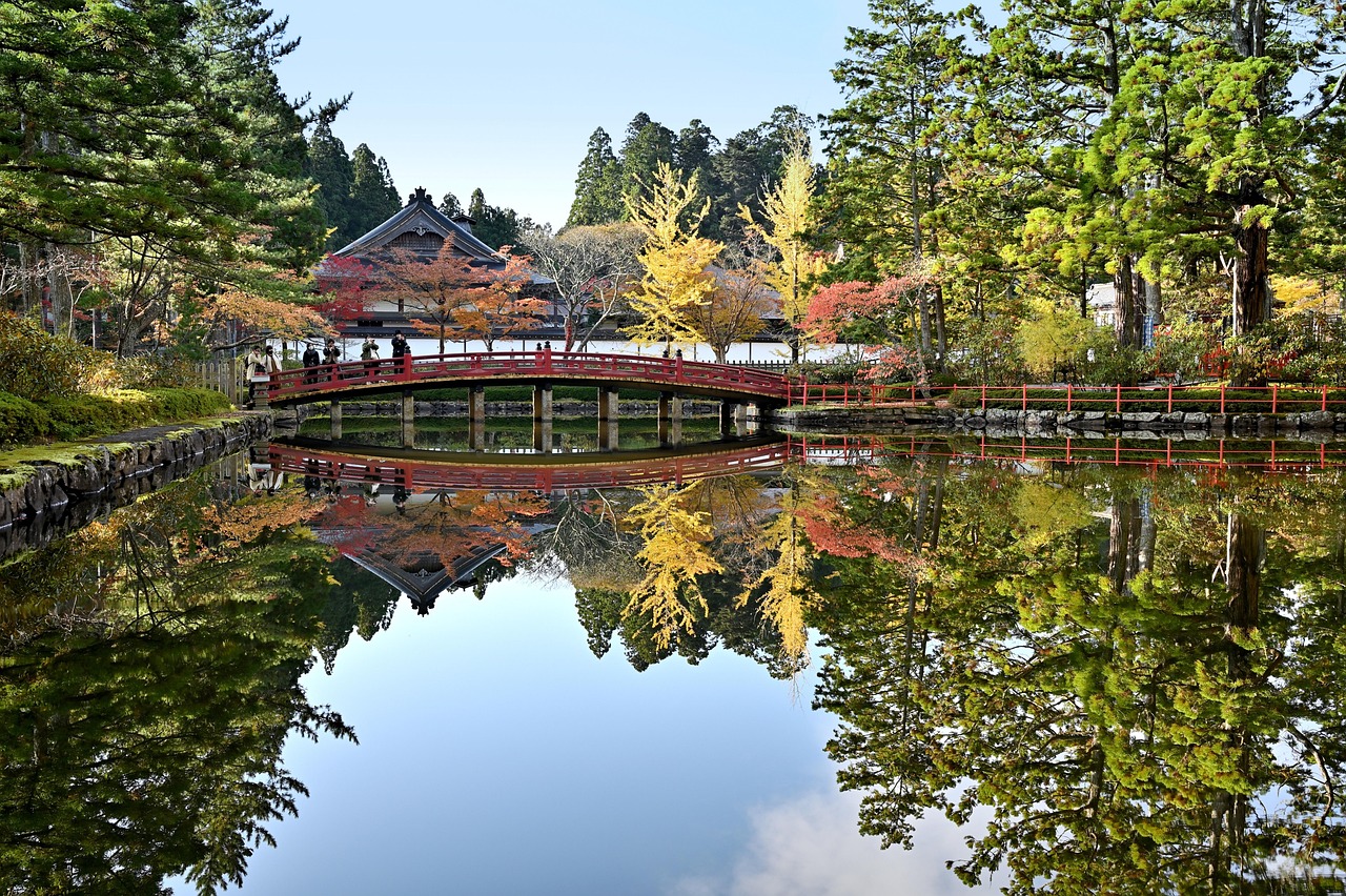 A still pond reflecting a red bridge and a temple hall among pines, cedars and autumn trees.
