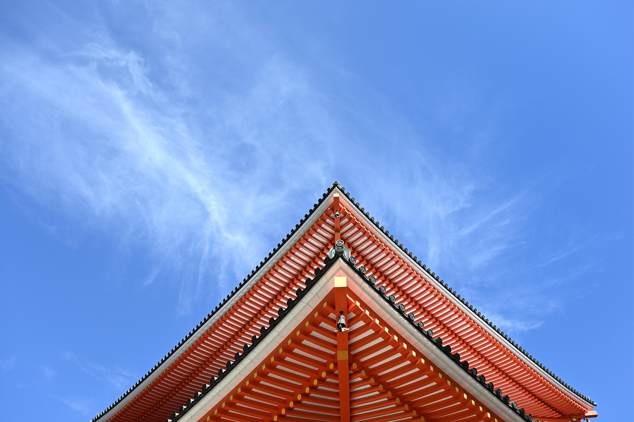 The upper roof of a vermilion pagoda against a blue sky with thin cloud.