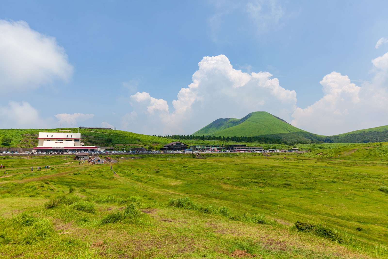 The Aso Volcano Museum beside a wide green grassland, a grass-covered volcanic hill and summer clouds behind