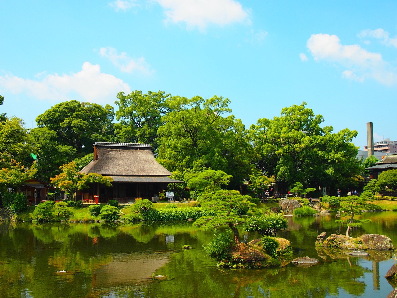A green pond with small rock islets and pines in Suizenji Jojuen, a thatched-roof building on the far bank