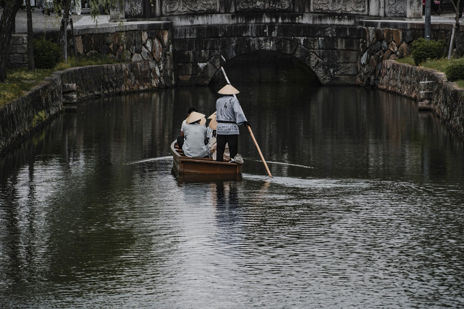 A Kurashiki river boat poled along the canal toward a stone bridge, the boatman and passengers in straw hats seen from behind