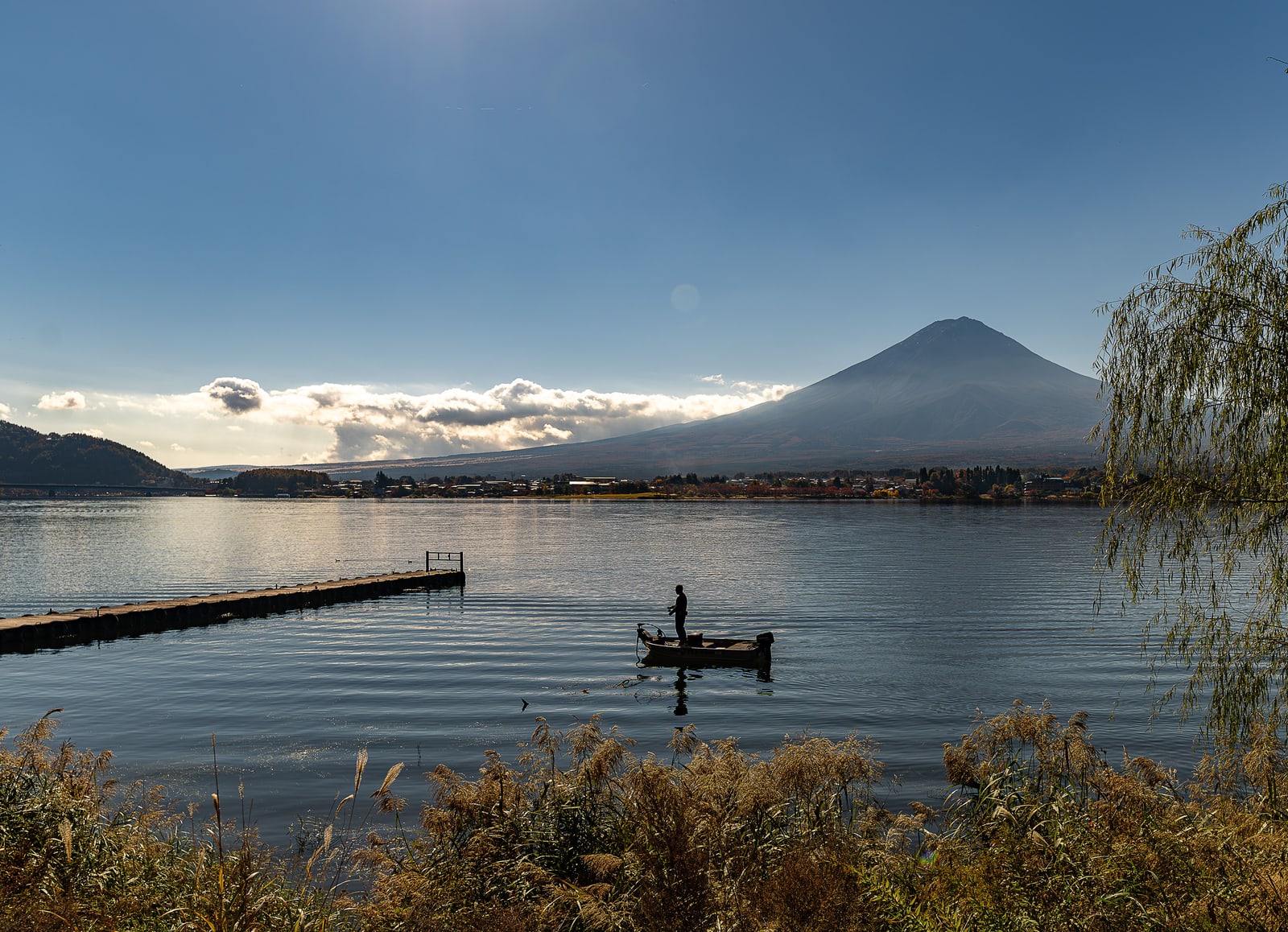 Mount Fuji without snow above Lake Kawaguchi, a band of cloud across its lower slopes, a long jetty and a small fishing boat on the water.