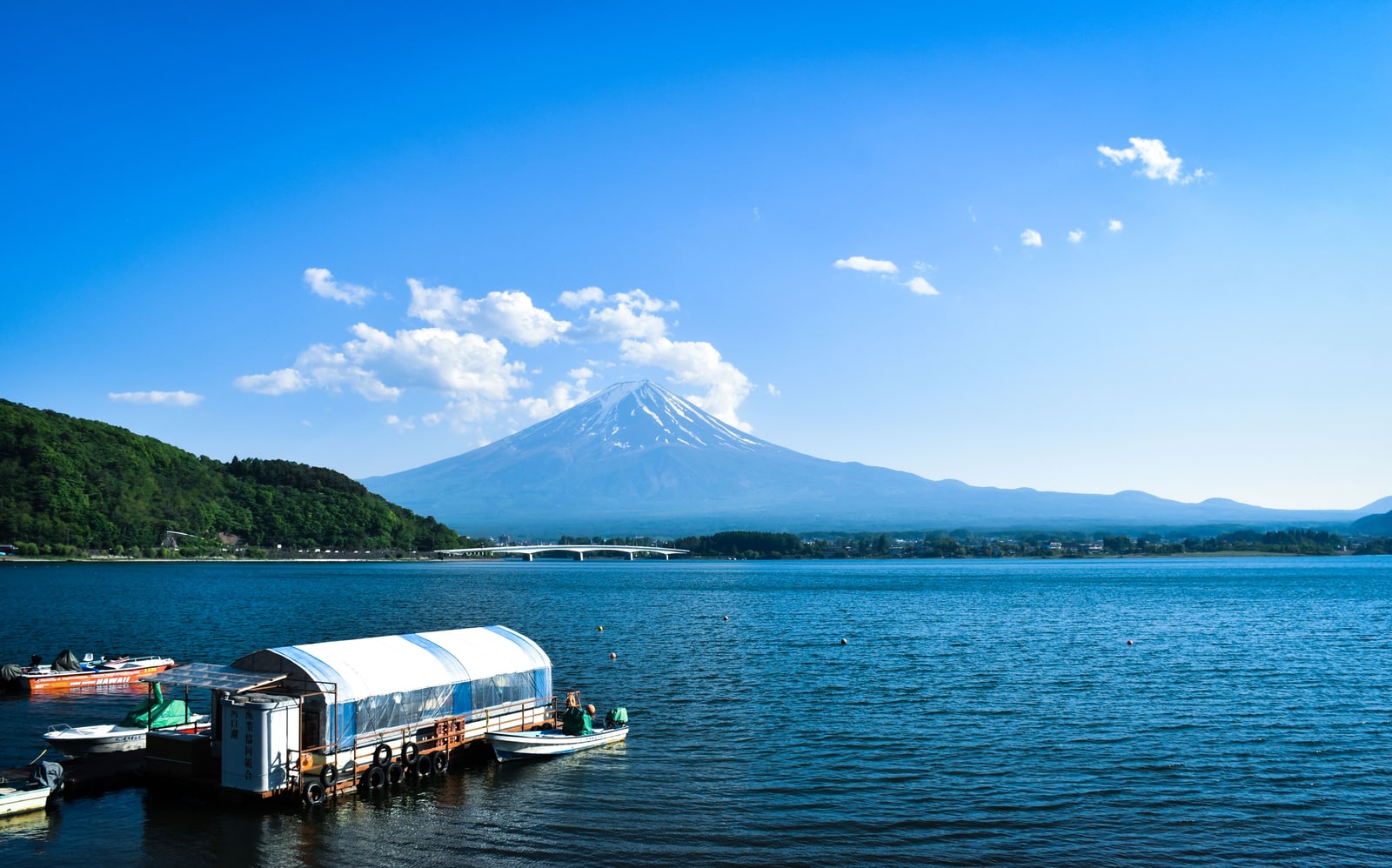 Snow-capped Mount Fuji under a deep blue sky with small clouds, moored boats at a jetty on Lake Kawaguchi in the foreground.