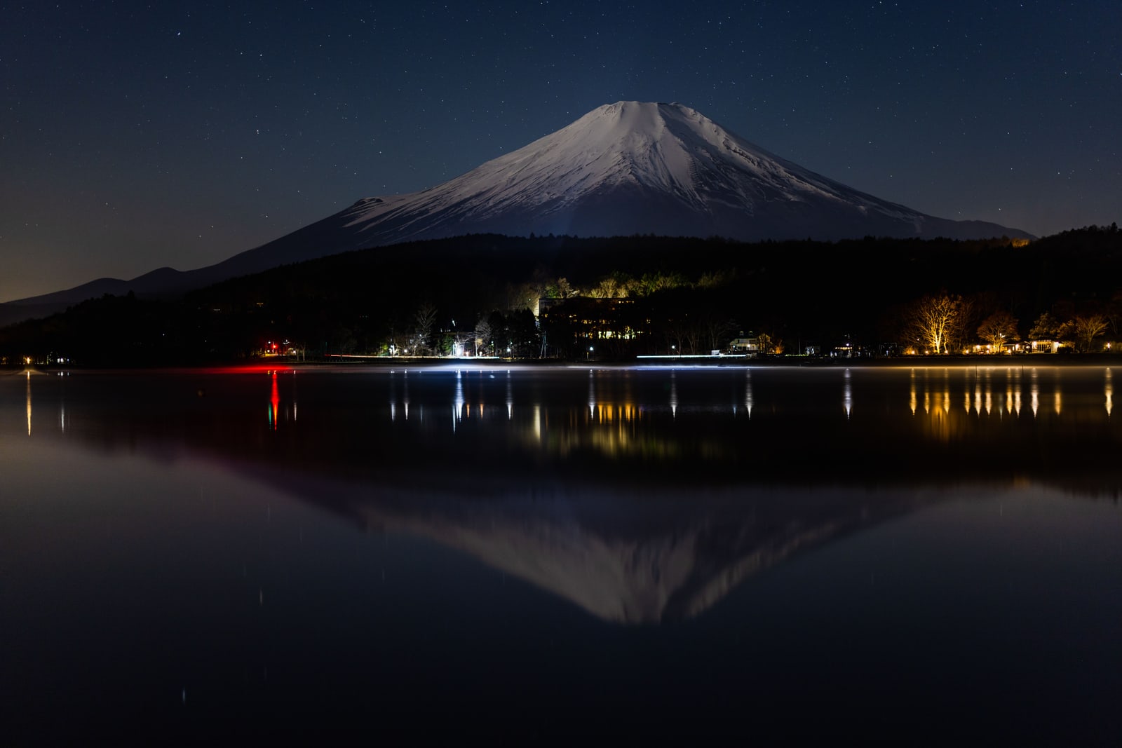 Mount Fuji at night, its snow faintly lit, the shoreline lights of Lake Kawaguchi reflected in black still water.