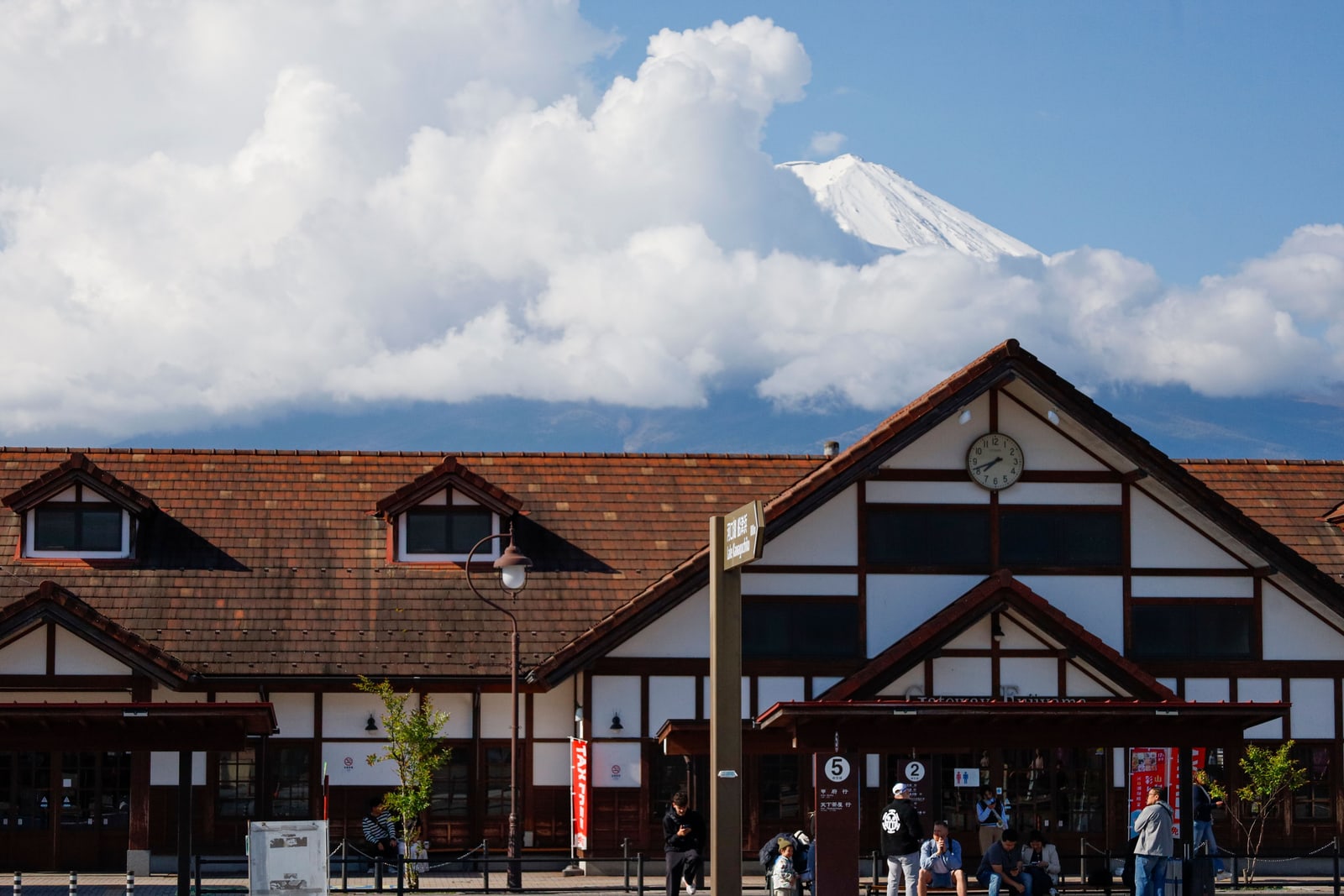 The timber-framed Kawaguchiko Station building with a clock on its gable, Mount Fuji mostly hidden behind a bank of white cloud.
