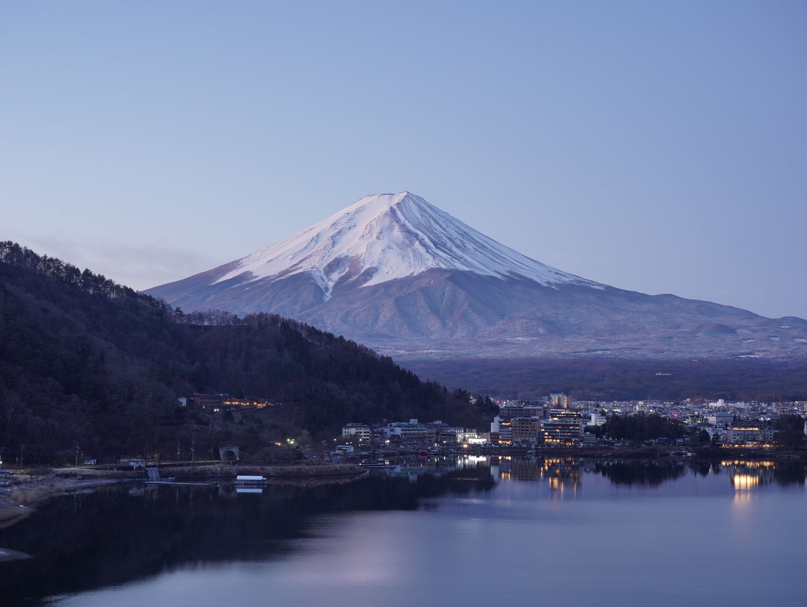Snow-capped Mount Fuji in blue twilight above a wooded ridge, the lights of the lakeside town at Kawaguchiko coming on along the water.