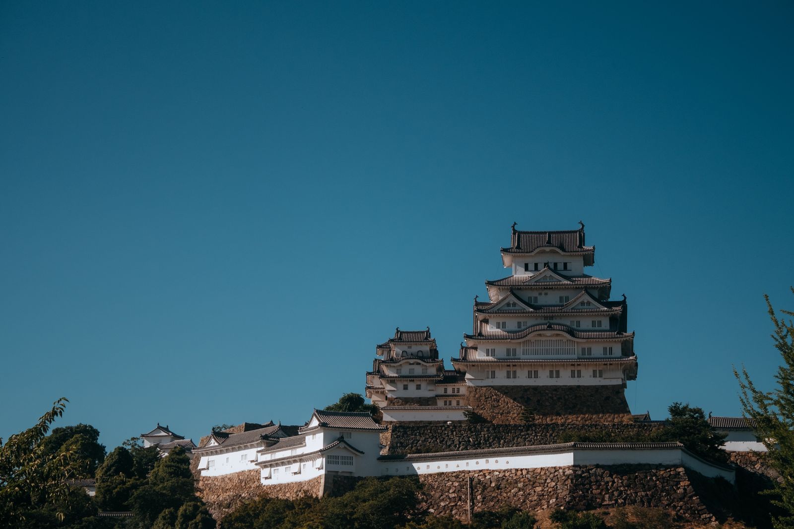 The white keep of Himeji Castle on its stone base against a deep unclouded sky, subsidiary turrets and the plastered wall running to the left.