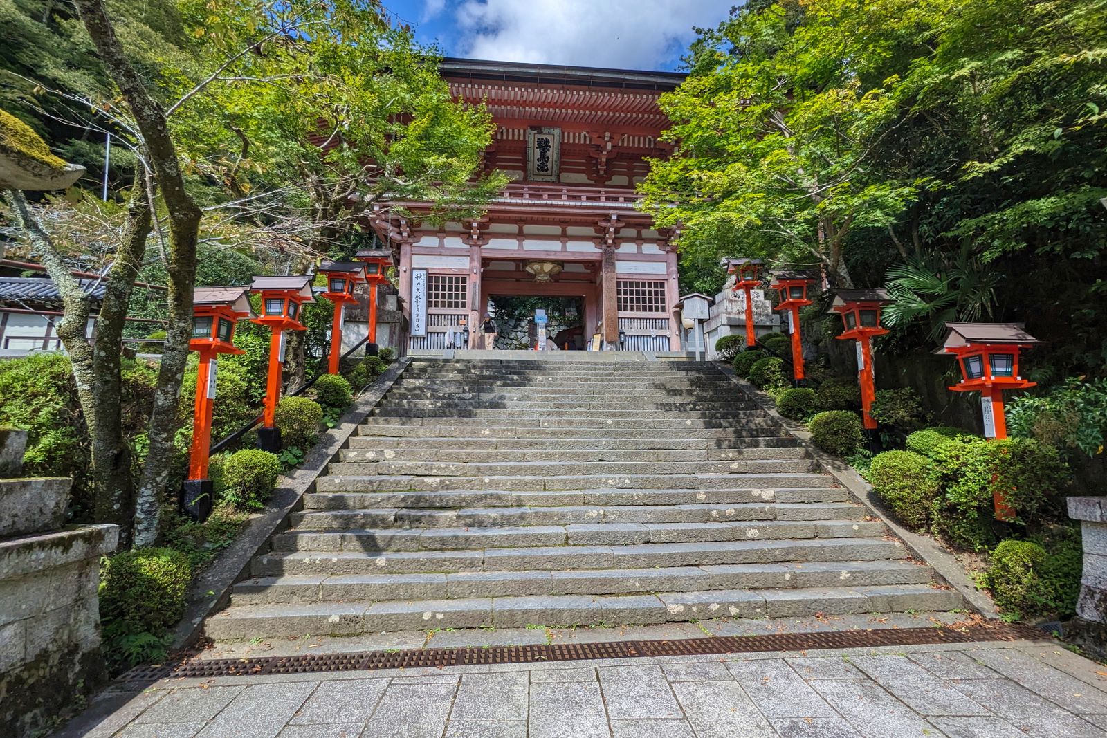 A broad flight of stone steps rising to a vermilion two-story gate flanked by two ranks of orange lantern posts, heavy green on both sides.