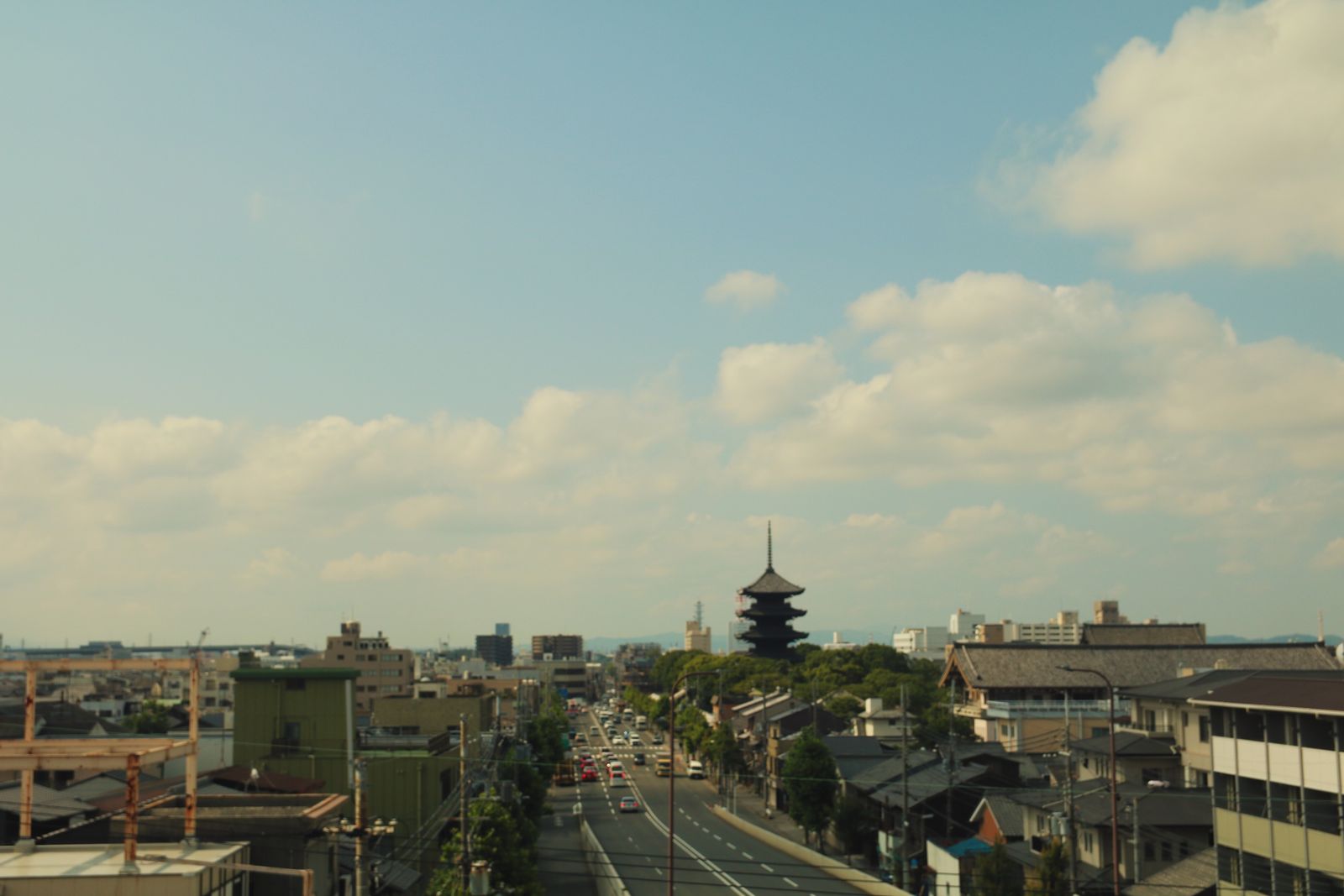 A raised view along a Kyoto avenue, low city roofs running to distant hills and a dark five-story pagoda standing above the rooflines.