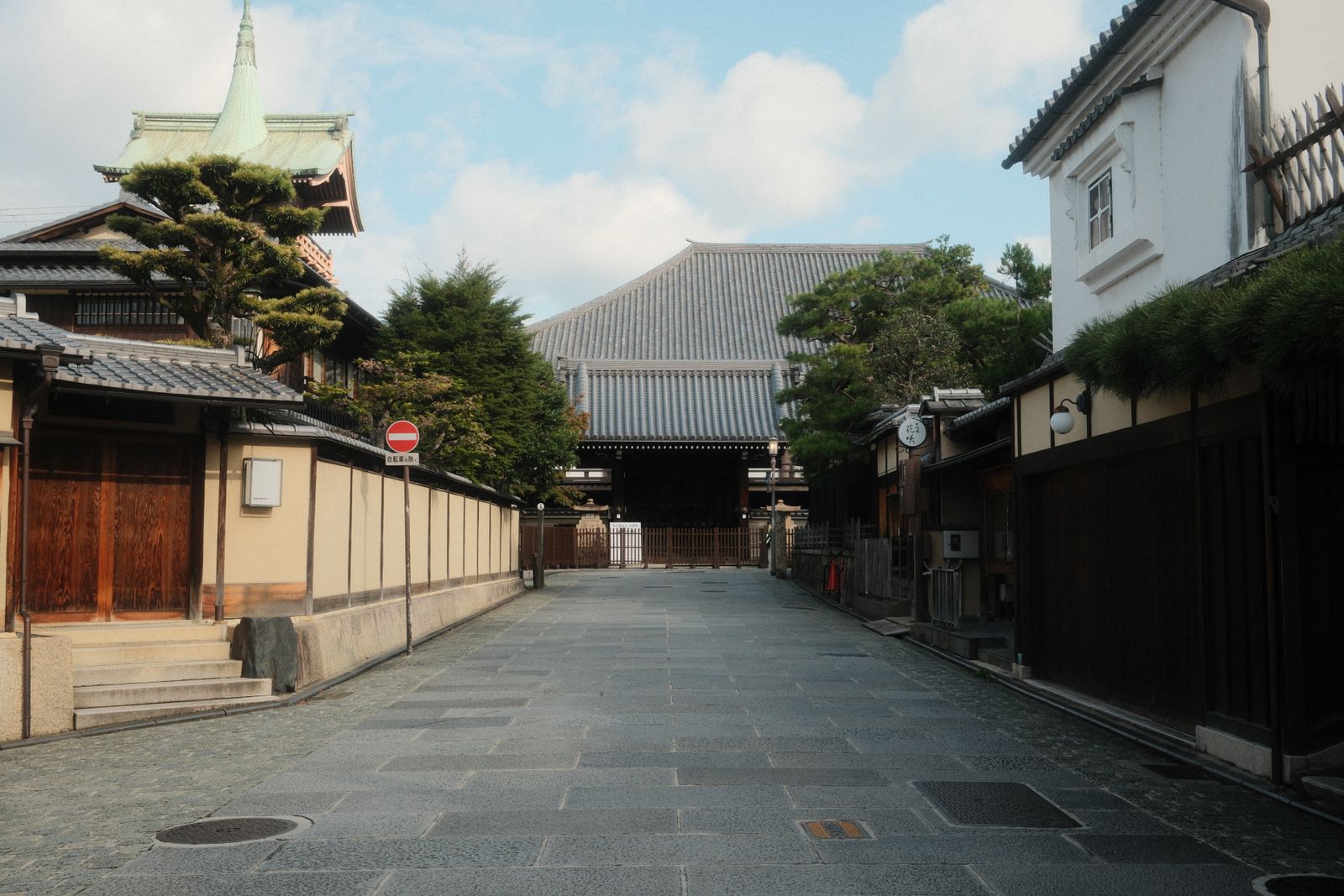 A wide stone-paved approach running to a temple gate in Kyoto, plastered walls and clipped pines at the left, machiya frontages at the right, nobody on it.