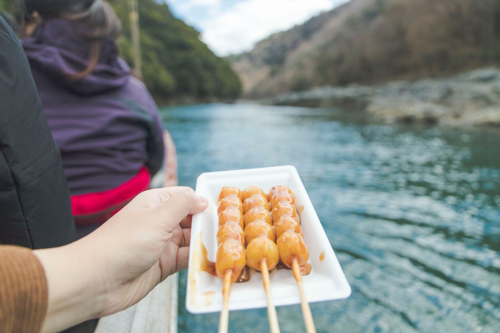 Three soy-glazed dango skewers on a tray, held aboard a boat on a Kyoto river, a passenger seen from behind.
