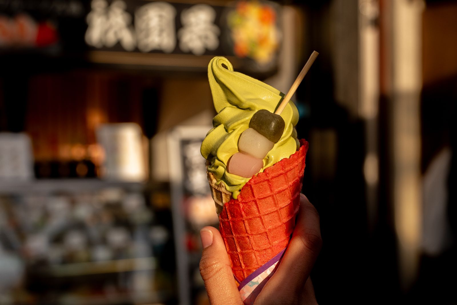 A hand holding a matcha soft-serve cone with a dango skewer on a Kyoto street, a sign blurred behind.