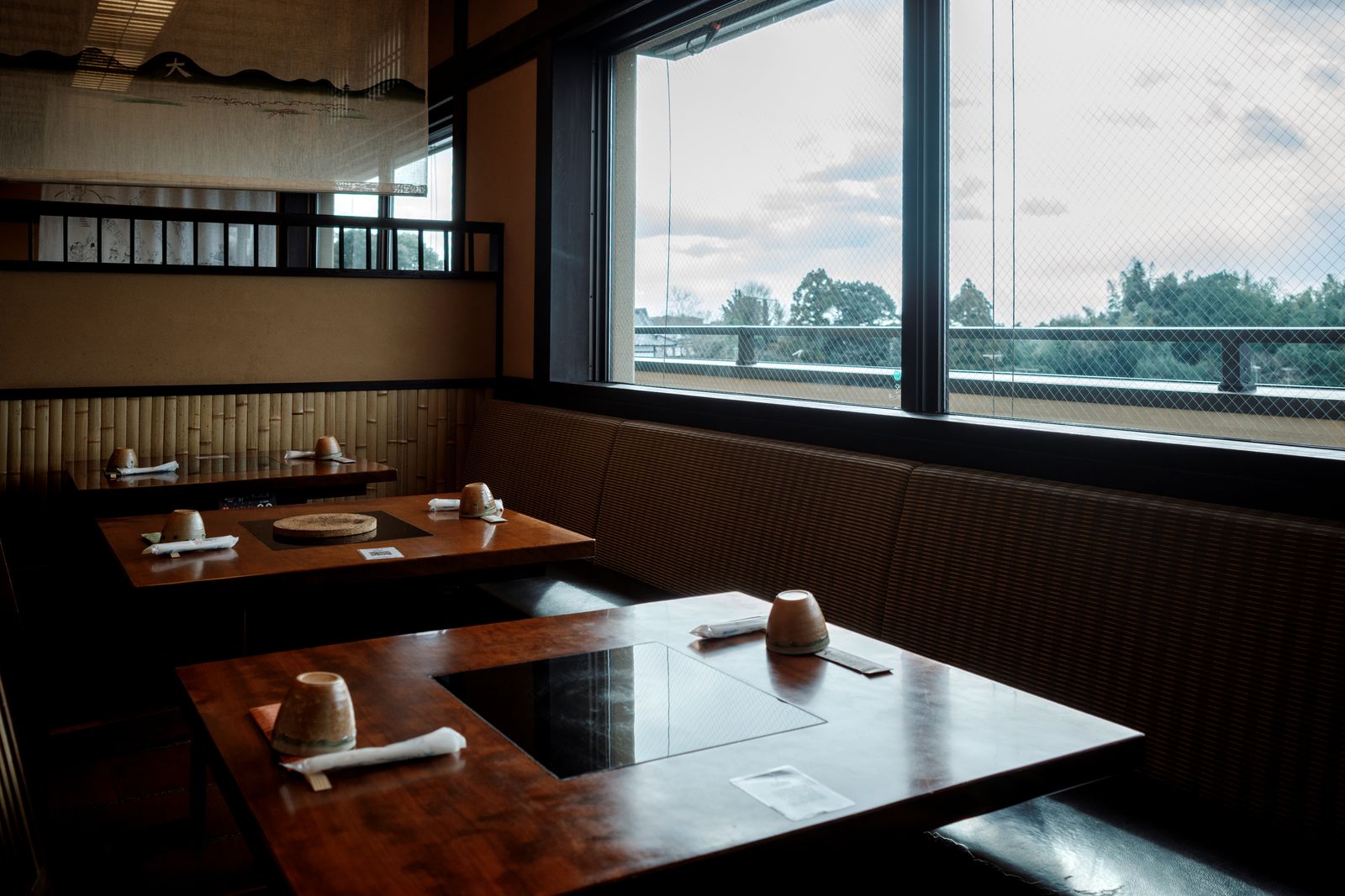 An empty restaurant dining room in Kyoto, tables with inset hot plates, bamboo wainscot, a window onto trees.