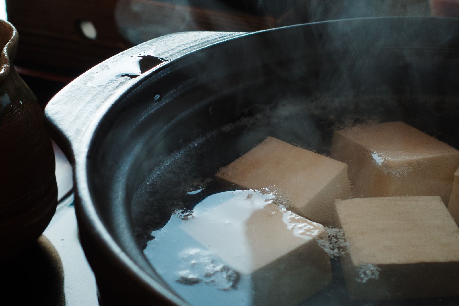 Cubes of tofu simmering in a dark glazed earthenware pot, steam rising, a small stoneware jug at the edge of a wooden table.