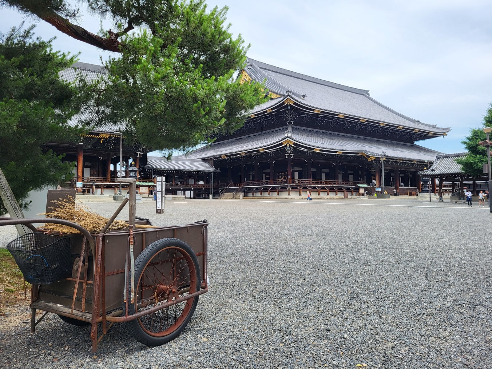 A large tiled temple hall behind a wide gravel courtyard, a pine branch and an old handcart with brooms in front