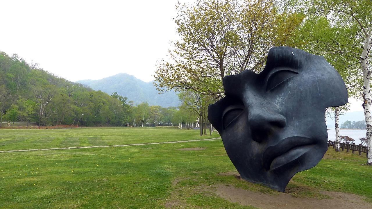 A large face sculpture on a lakeside lawn by Lake Toya, Hokkaido, birch trees and forested hills behind