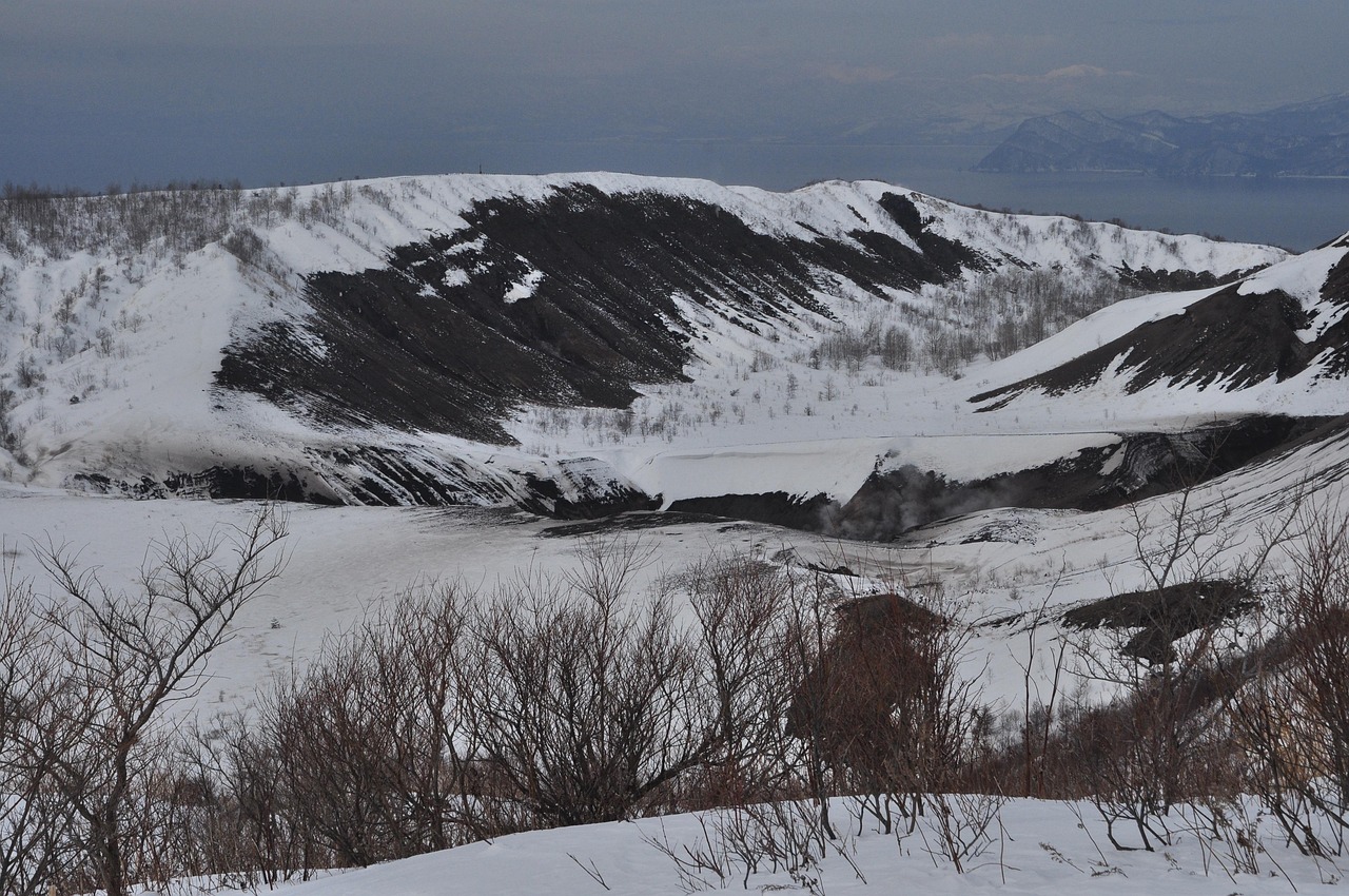 A snow-covered volcanic crater with steam rising, water and hills beyond, Hokkaido