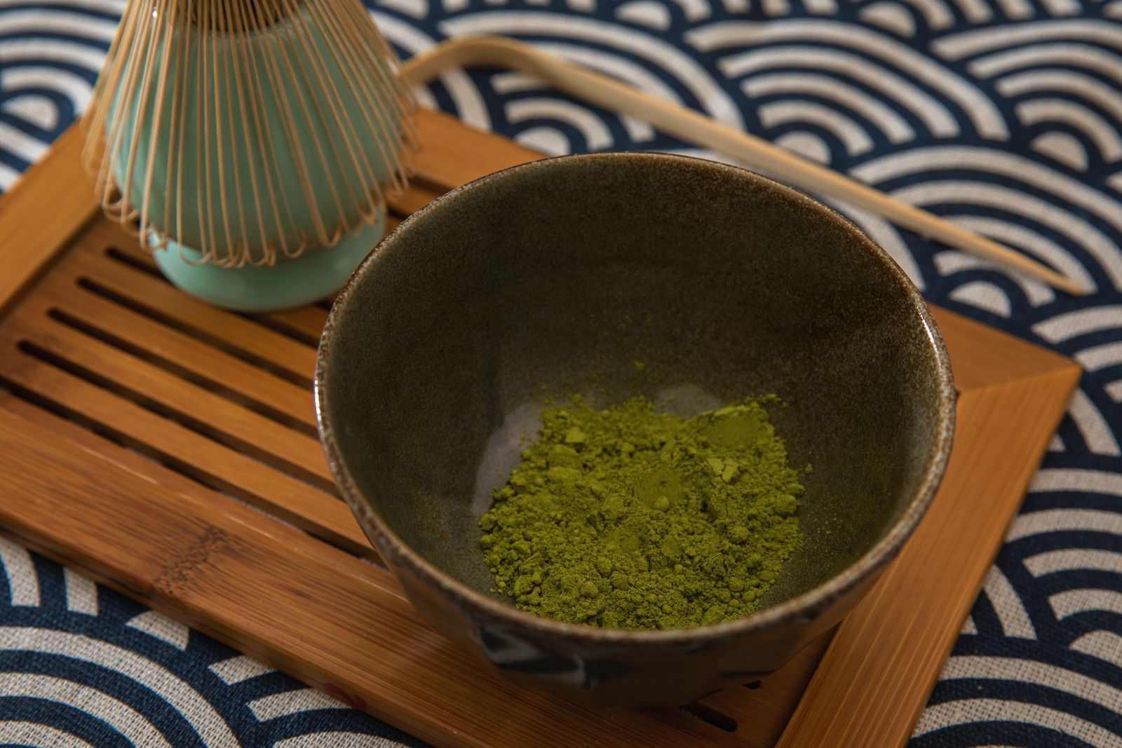 A tea bowl of un-whisked matcha powder beside a bamboo whisk on its holder and a bamboo scoop on a tray, on patterned cloth.