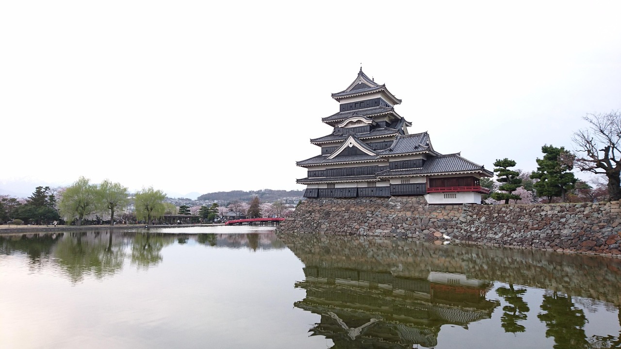 The black-and-white keep of Matsumoto Castle reflected in its moat, cherry trees in blossom at left