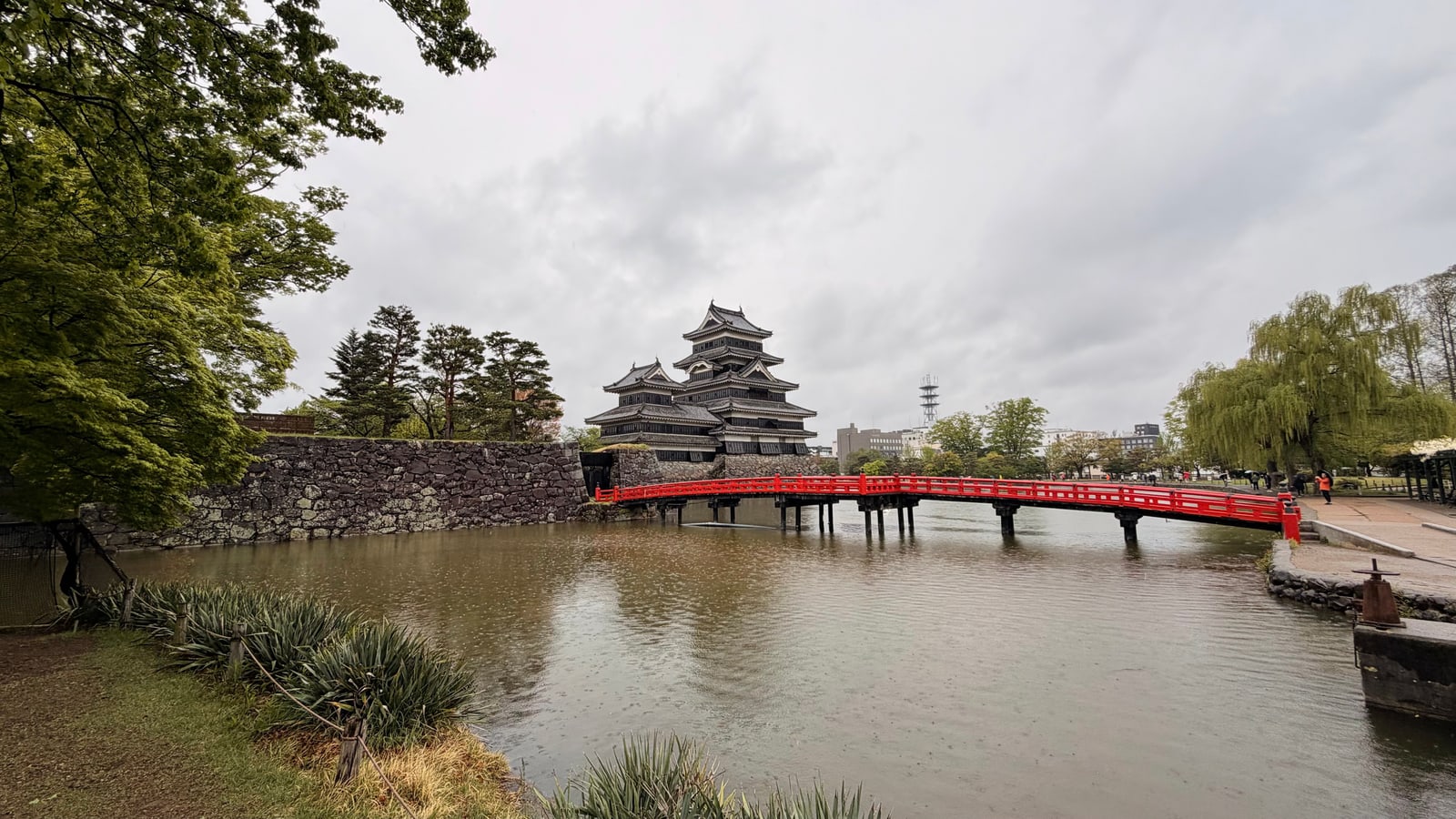 Matsumoto Castle beyond its moat on an overcast spring day, a red wooden bridge crossing the water at right