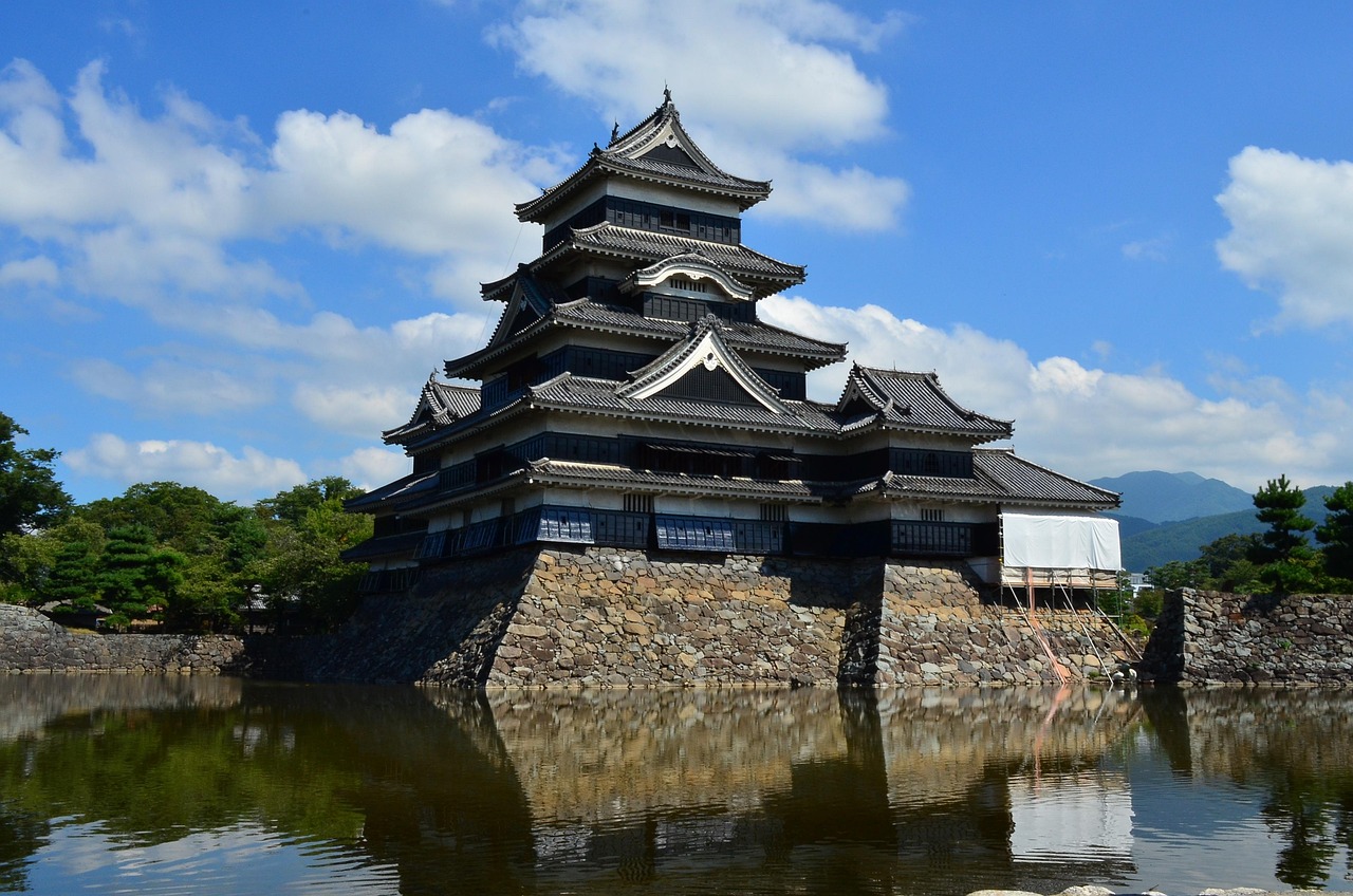 Matsumoto Castle and its turrets reflected in the moat under a blue summer sky, one corner under scaffolding