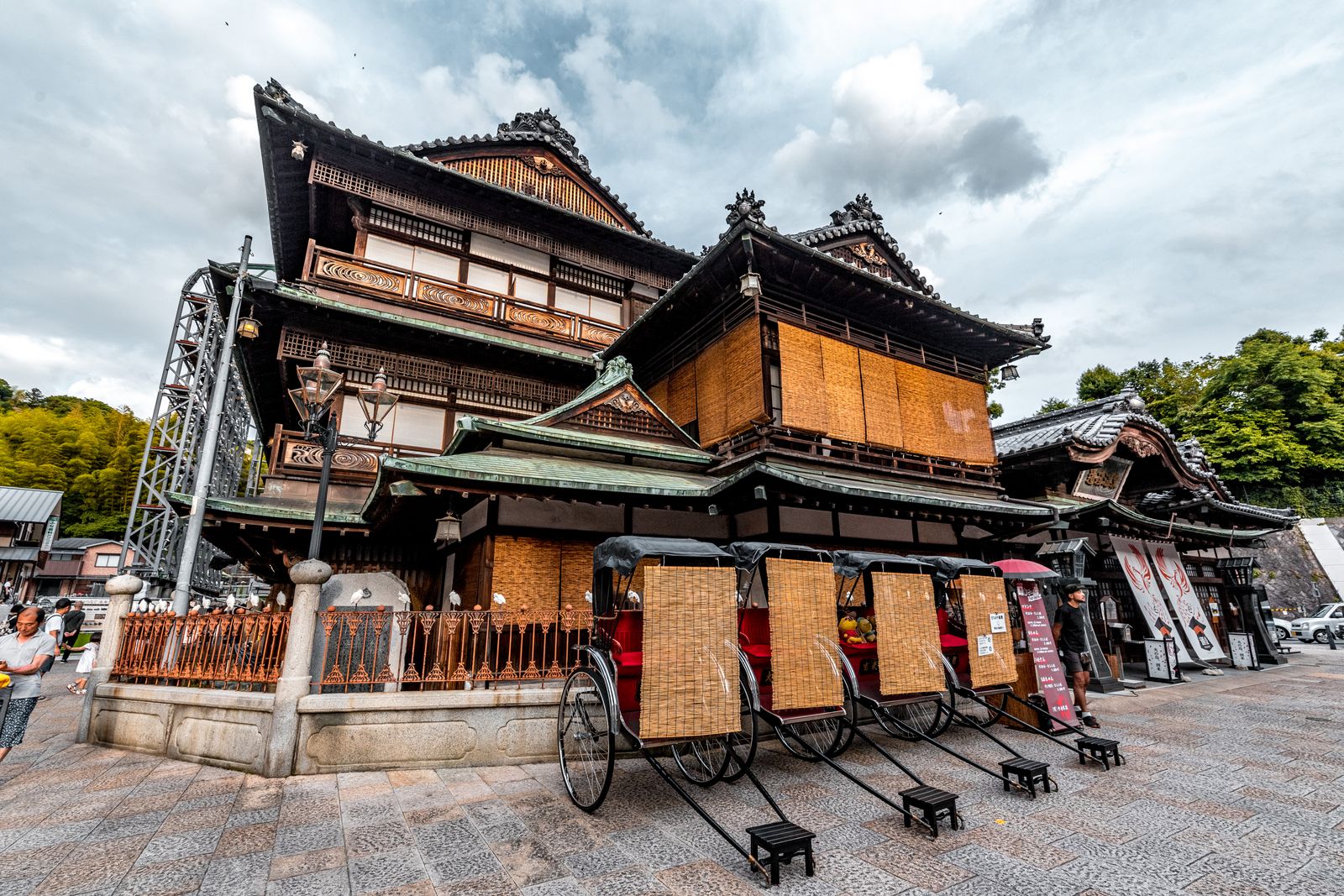 The wooden Dogo Onsen Honkan bathhouse with rickshaws parked in front under a cloudy sky