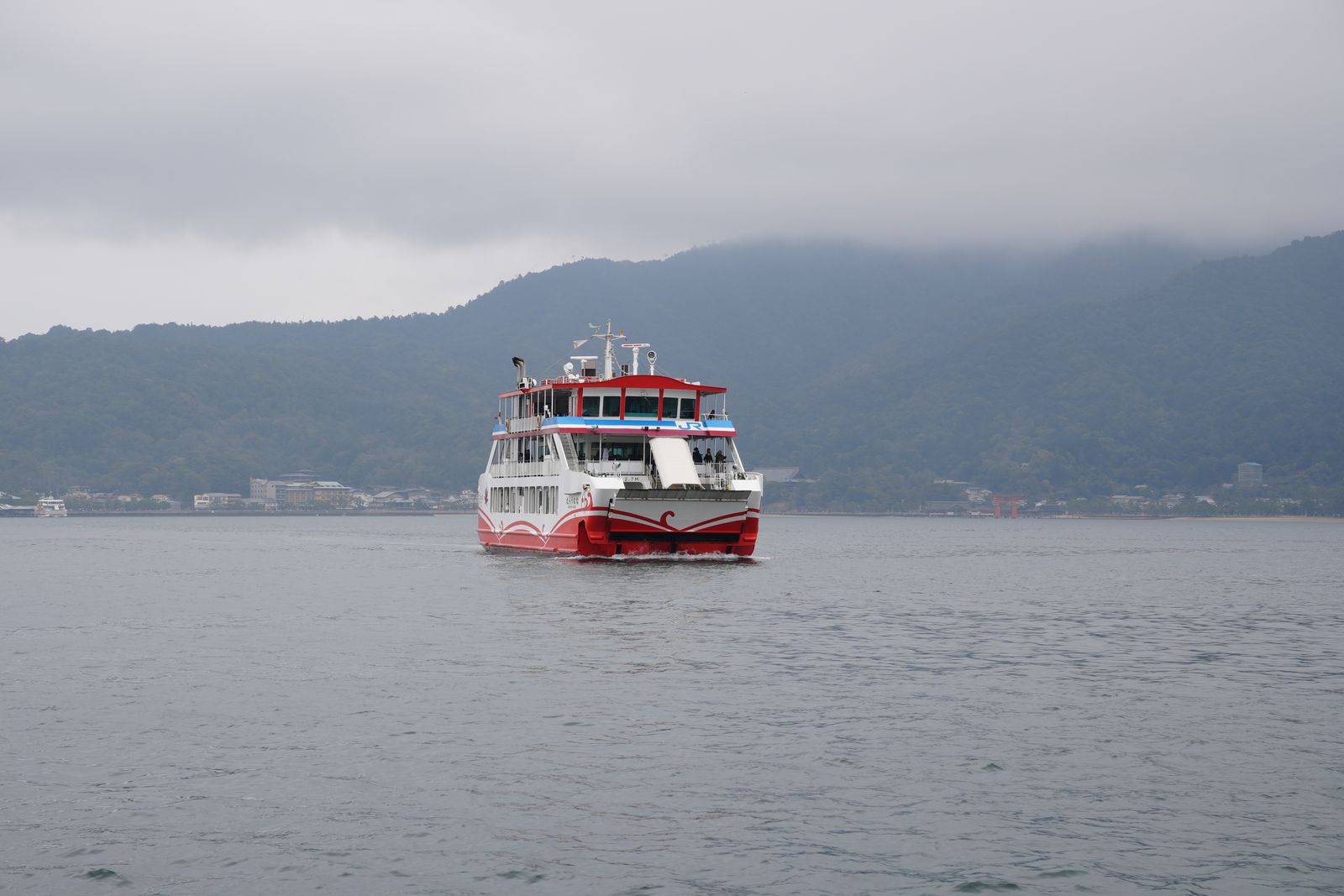 A JR ferry on the water off Miyajima on an overcast day, the island's forested hills behind