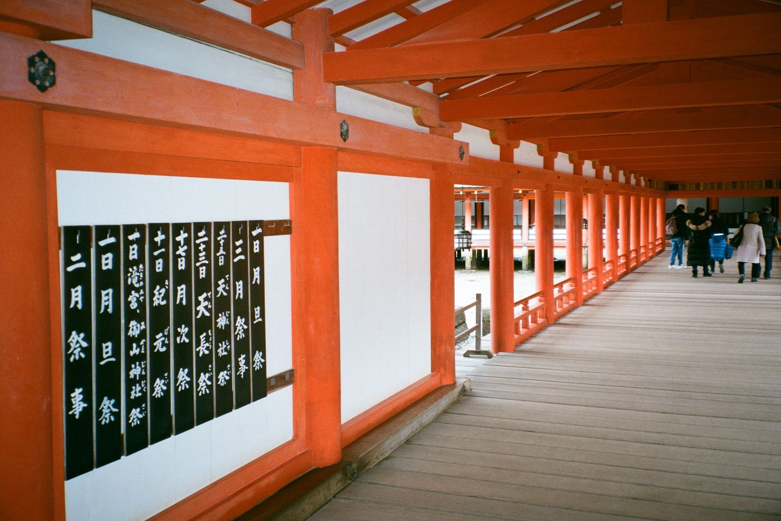 A vermilion corridor of Itsukushima Shrine with festival plaques on the wall and the tide out between the pillars, visitors walking away