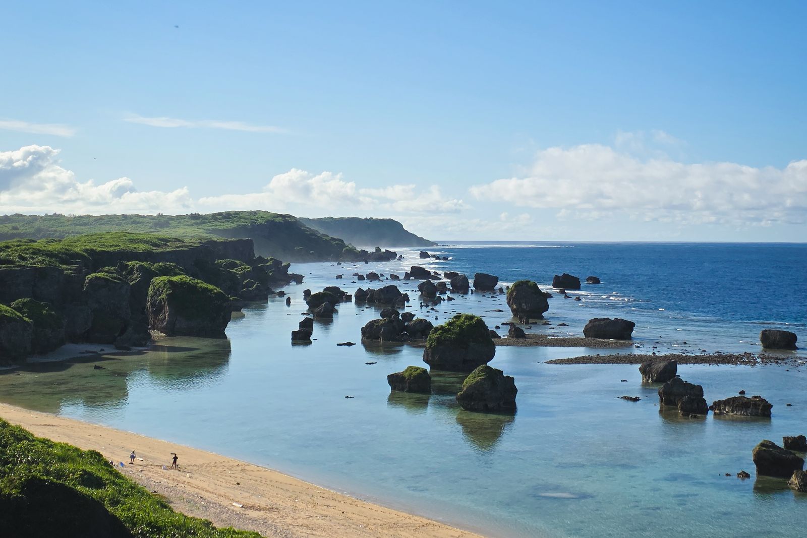 A rocky coast on Miyakojima with moss-covered boulders standing in shallow water beside a sand beach