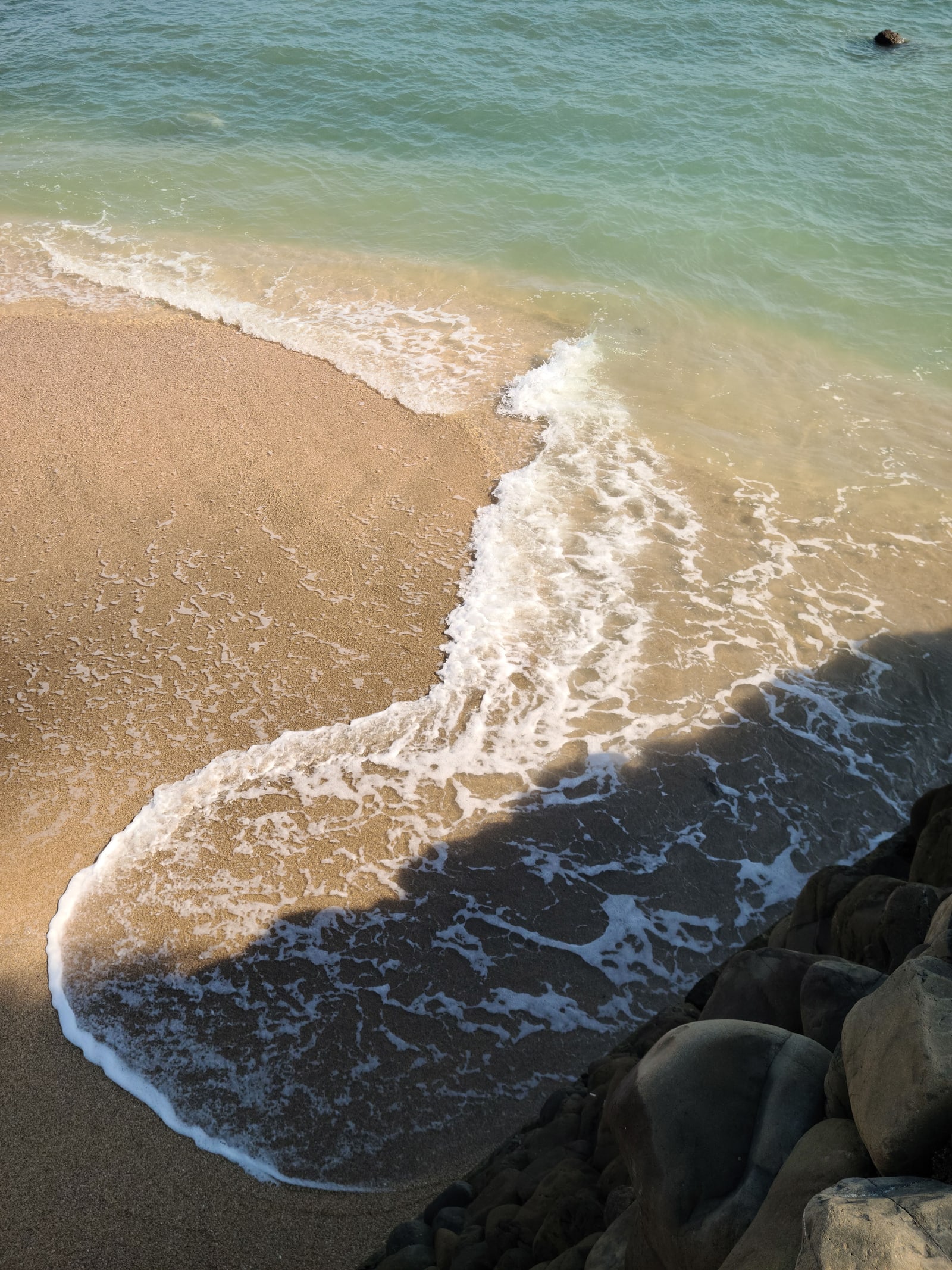 A wave running up a sandy shore beside dark rocks