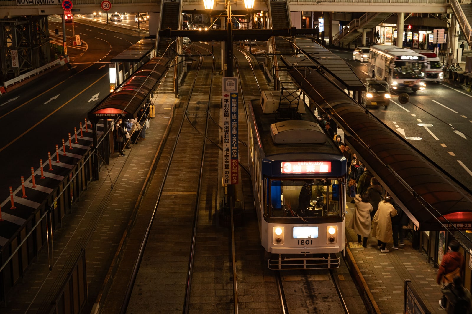 A tram marked 3 at a tram stop at night, passengers waiting under the shelter roofs, a bus and cars on the road and a signpost listing destinations in Japanese