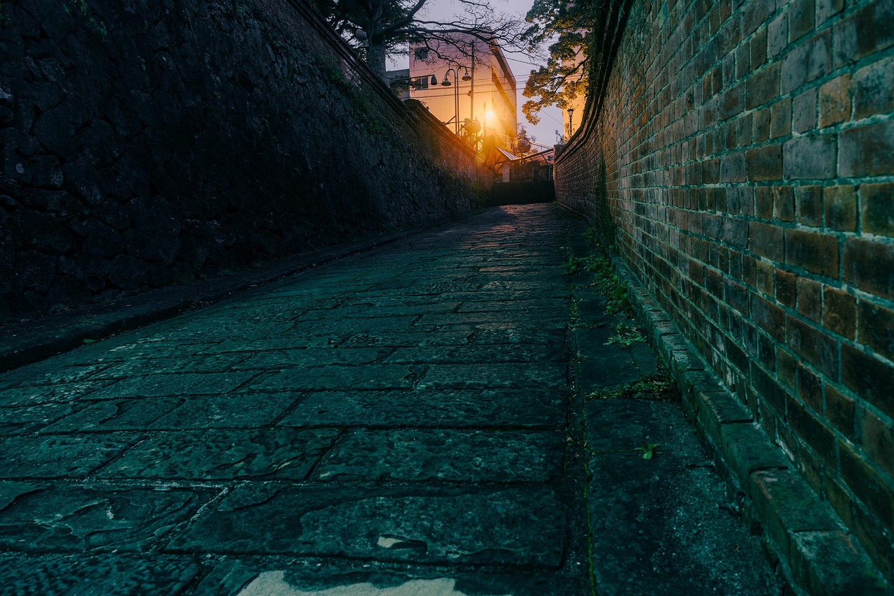 A stone-paved slope climbing between a dark rough stone wall on the left and a weathered brick wall on the right, with street lamps at the top in dusk light