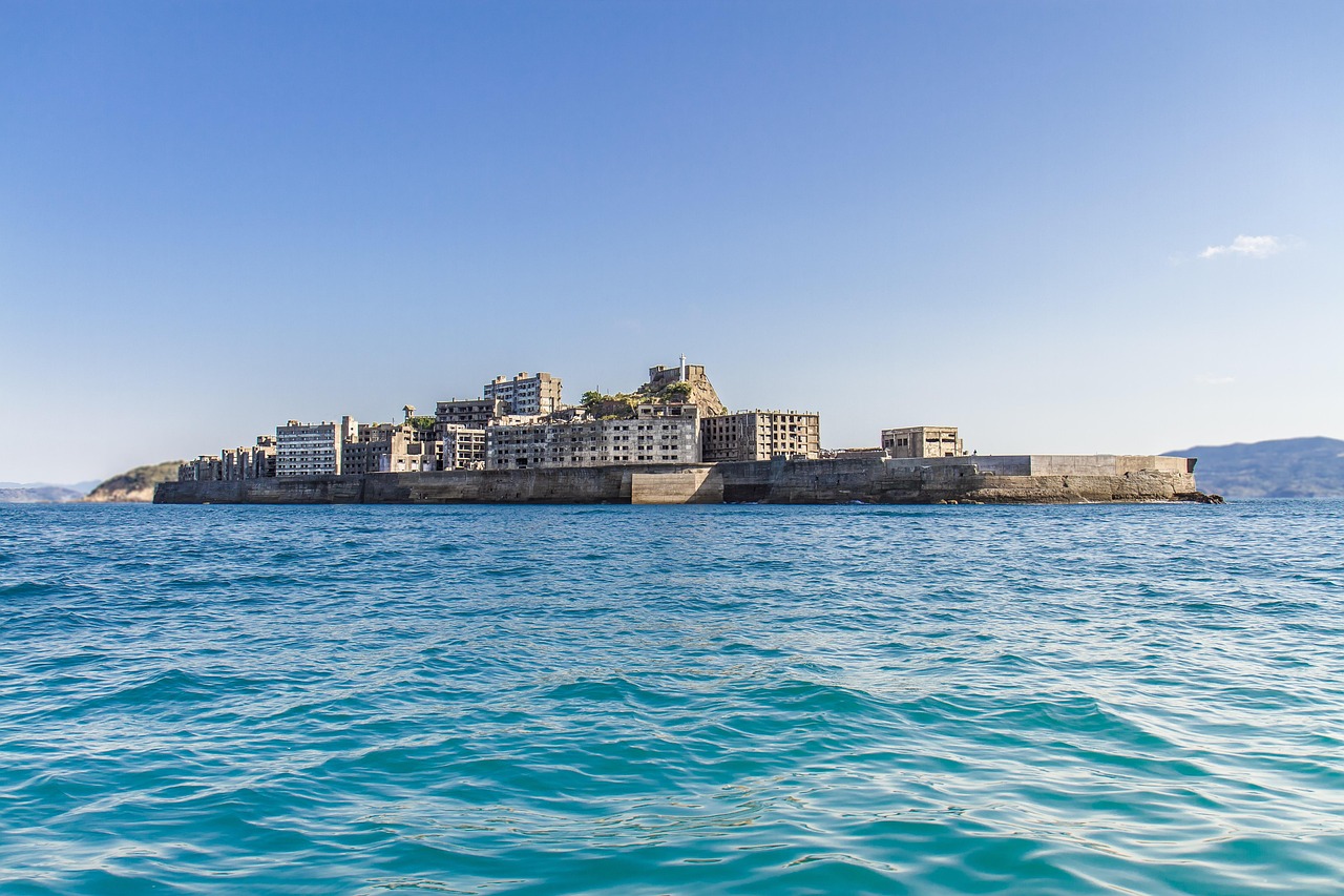 A low island of ruined concrete buildings behind a sea wall, seen from the water under a clear blue sky with turquoise sea in front