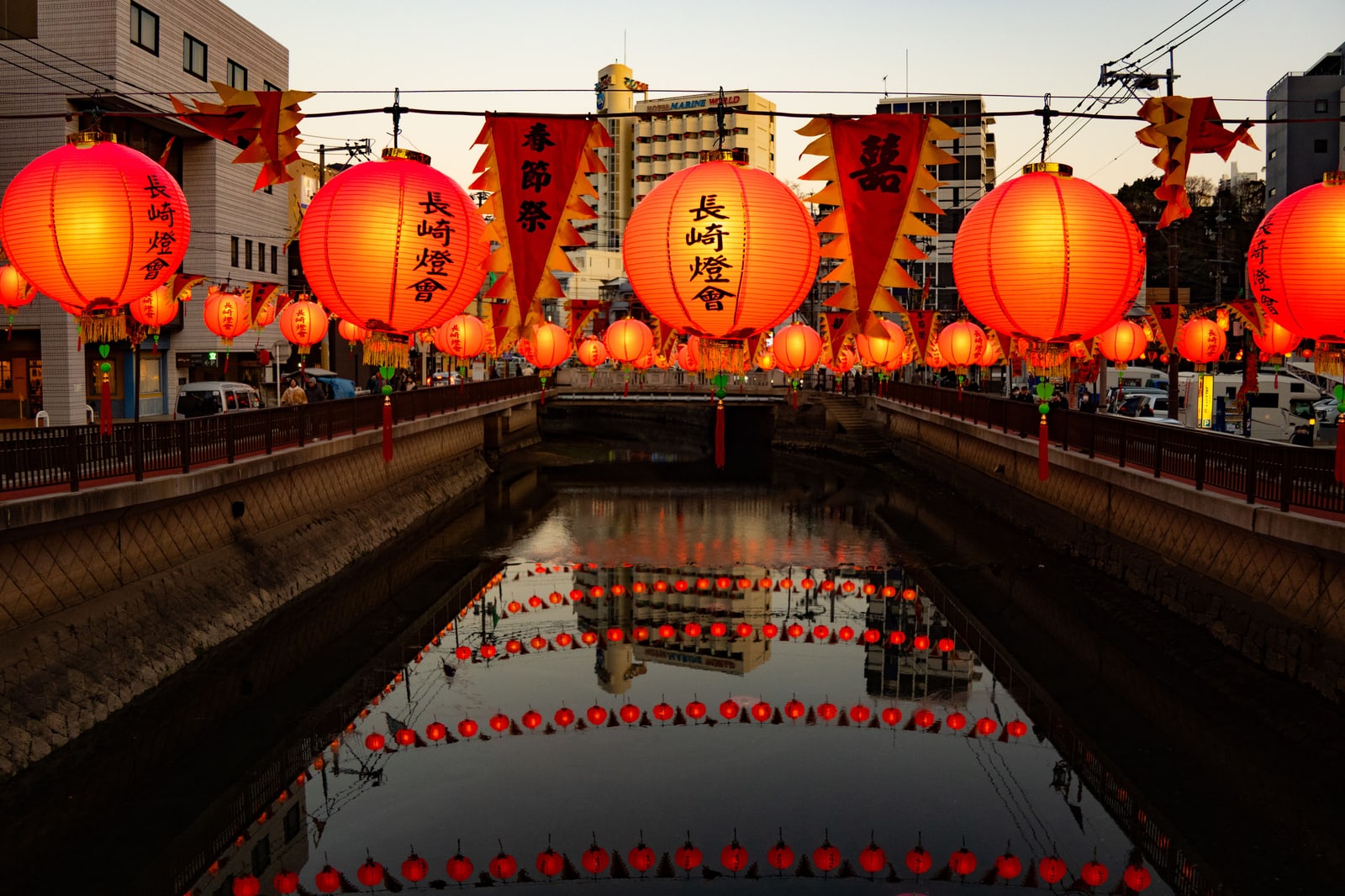 Rows of red paper lanterns printed with 長崎燈會 hung over a canal, their reflections in the still water, with buildings and a hotel sign beyond at dusk