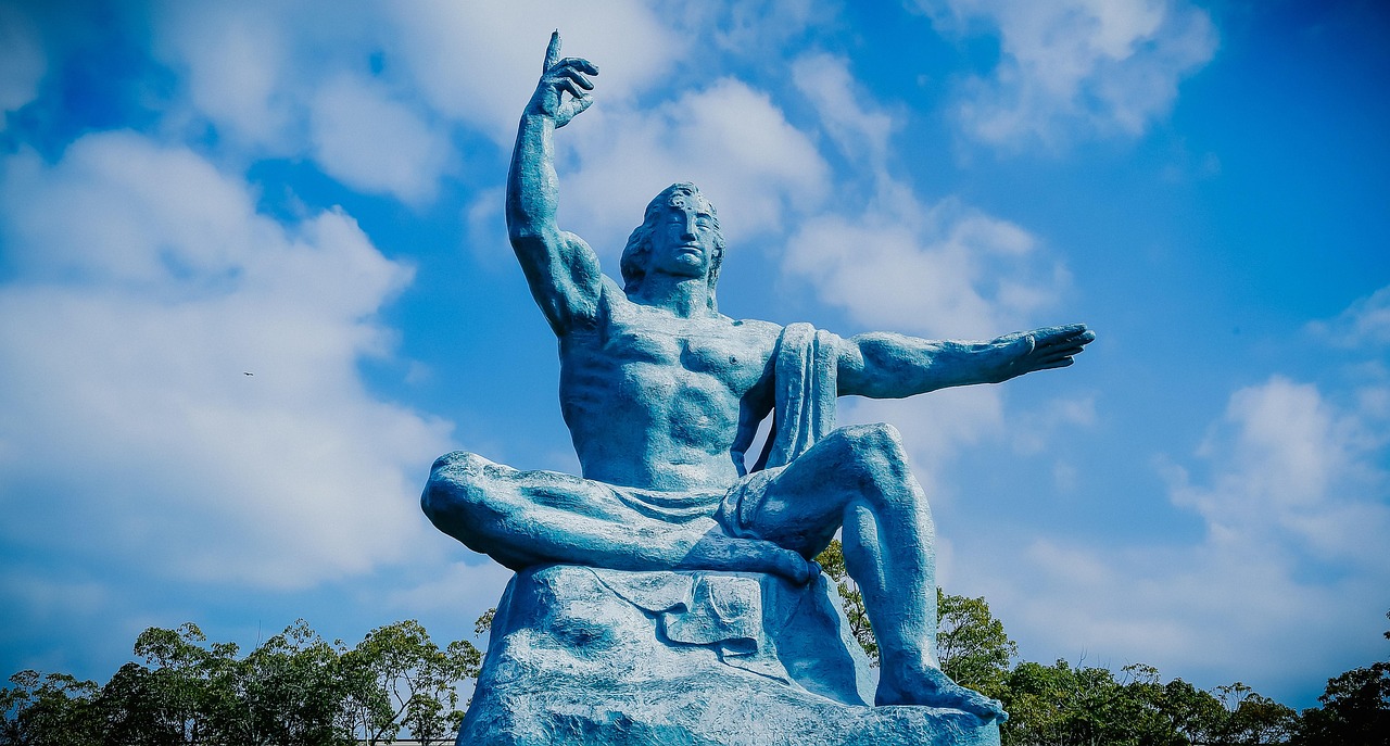 A blue-grey statue of a seated figure with the right arm raised and the left arm stretched out sideways, against a blue sky with white clouds