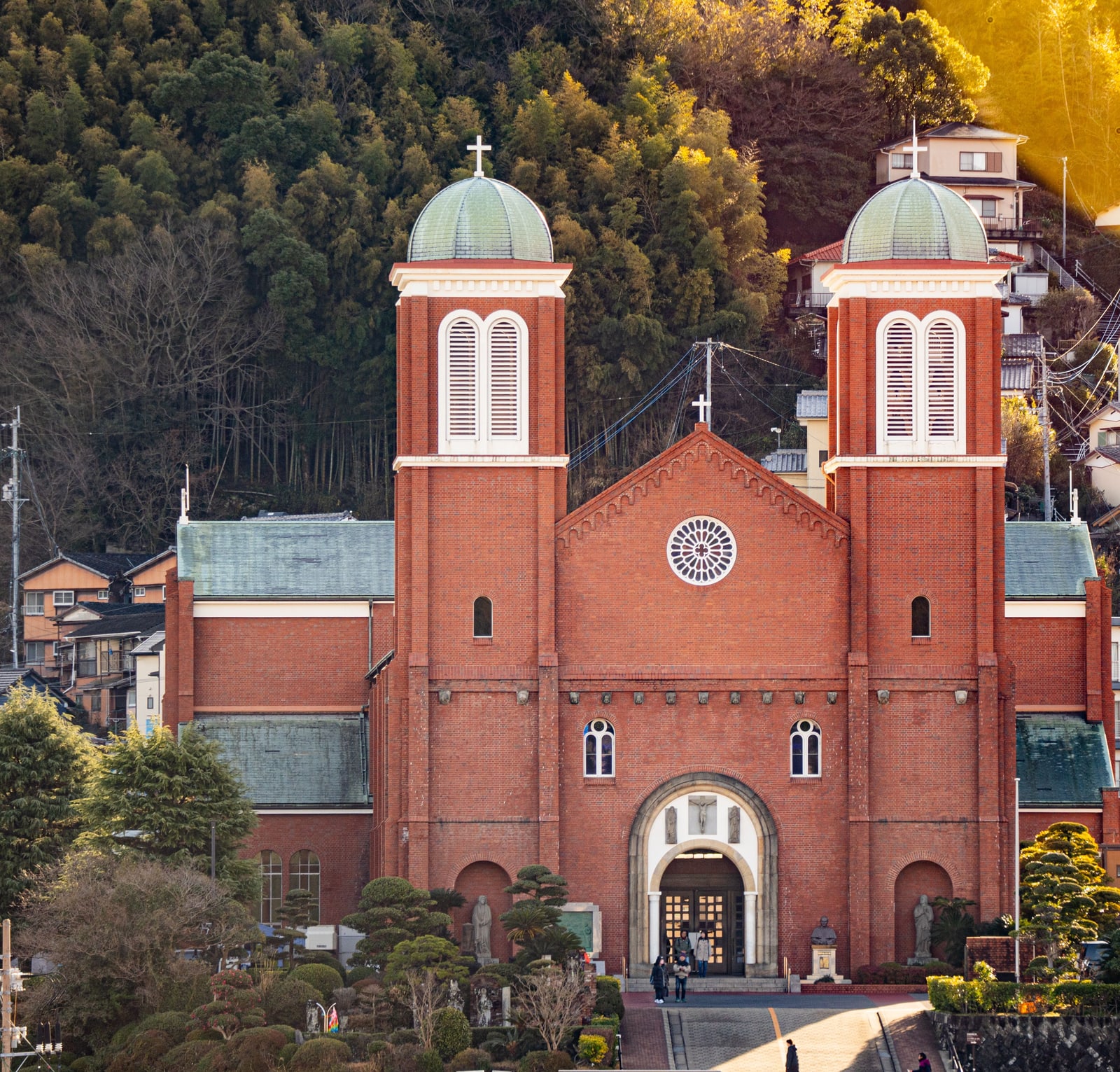The red-brick front of a church with two towers topped by green domes, a round rose window and an arched entrance, with a wooded hillside behind